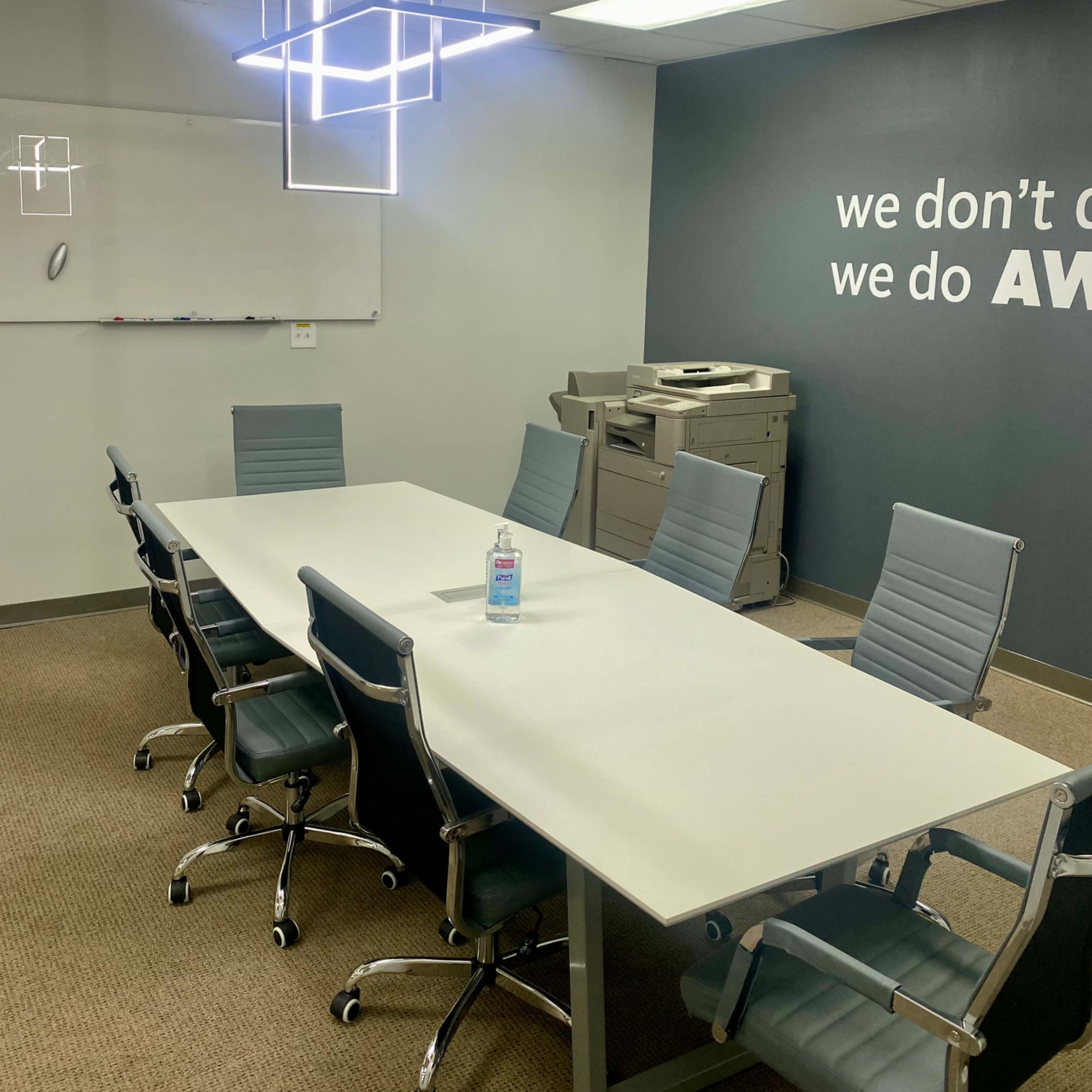 A conference room features a long table surrounded by chairs, with a modern light fixture overhead and a whiteboard on one wall.