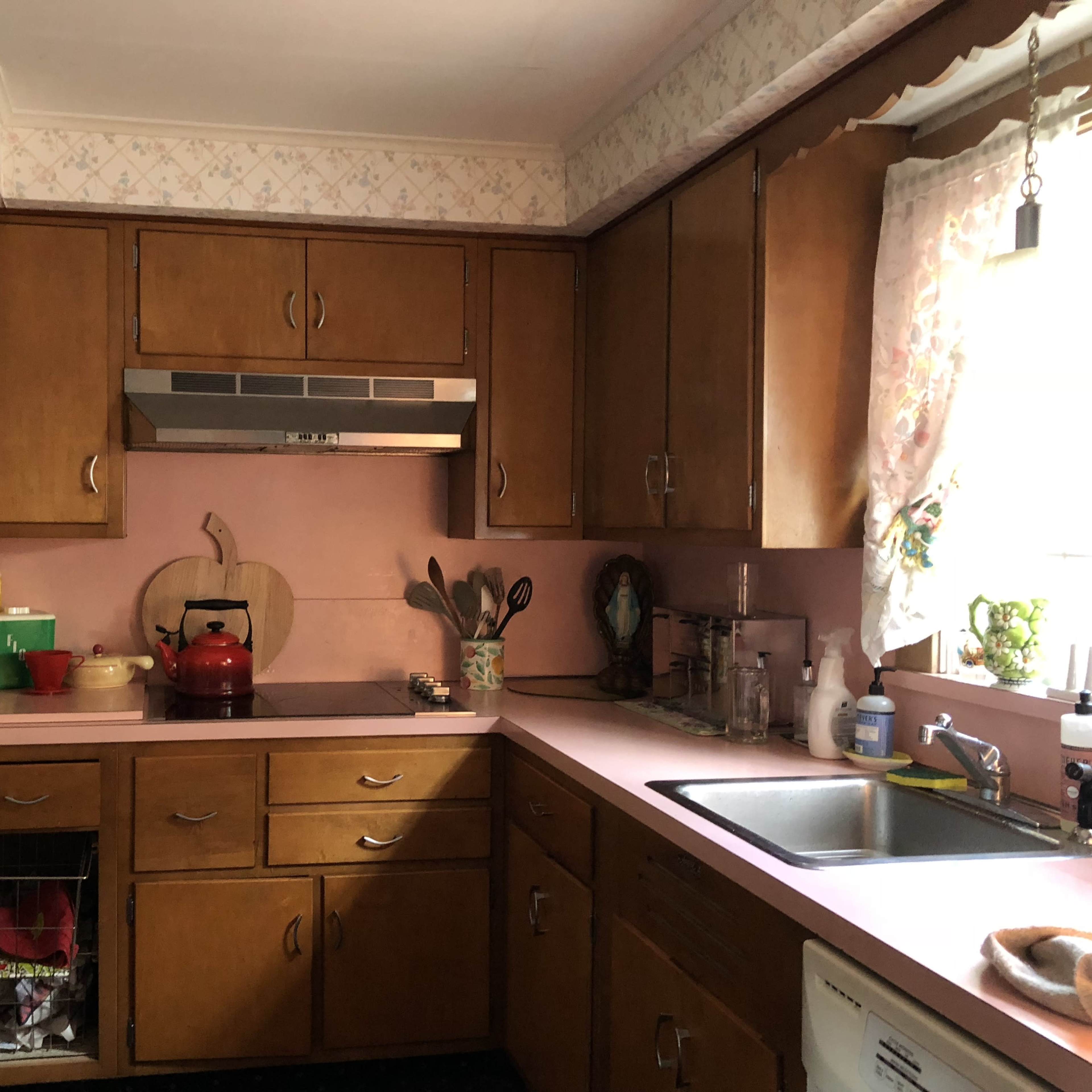 A kitchen with wooden cabinets, a pink countertop, and an assortment of cooking items including a kettle and utensils near a sink and stove.