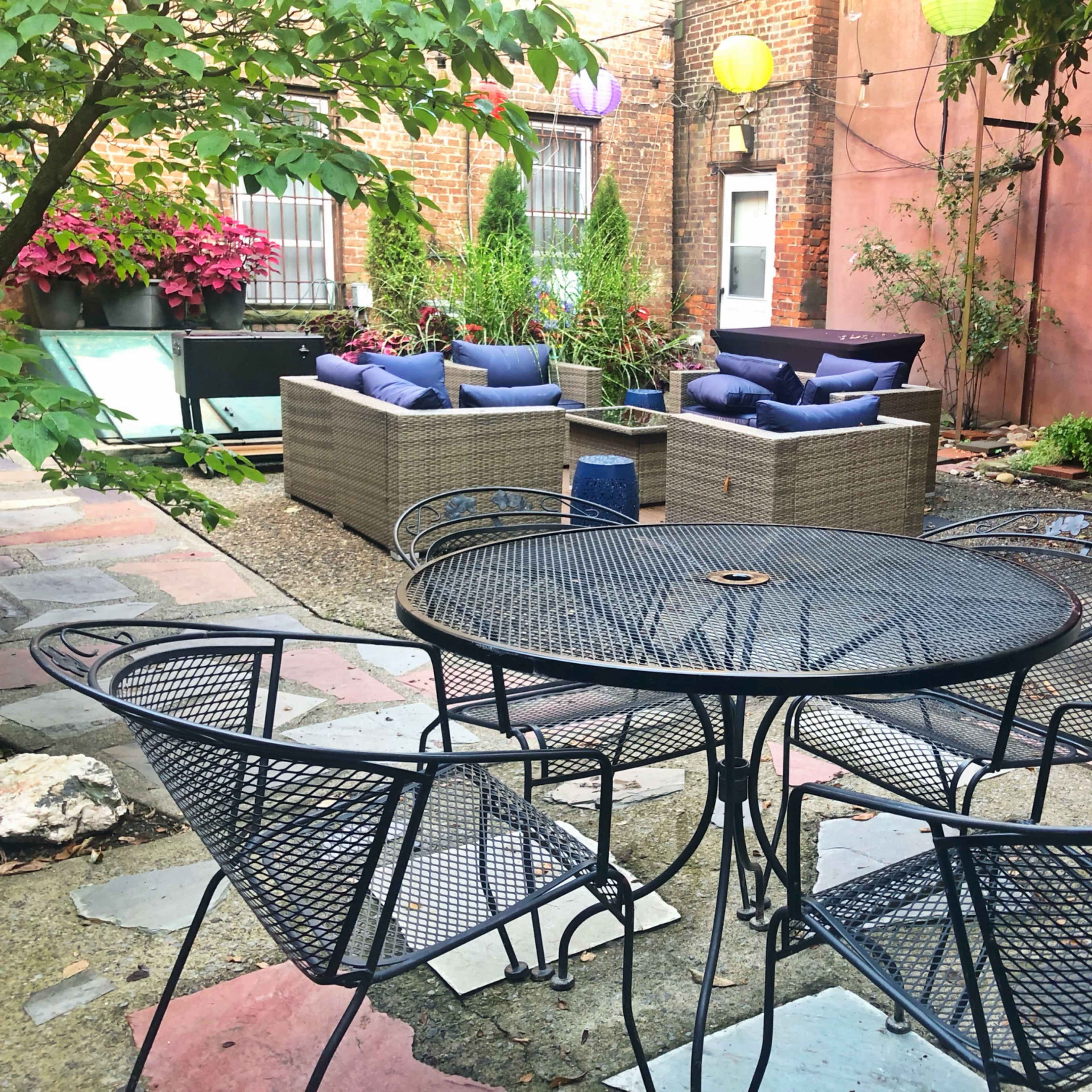 A small outdoor seating area features a round black metal table surrounded by four wire chairs, with two couch seating arrangements in the background amidst potted plants and decorative lanterns.