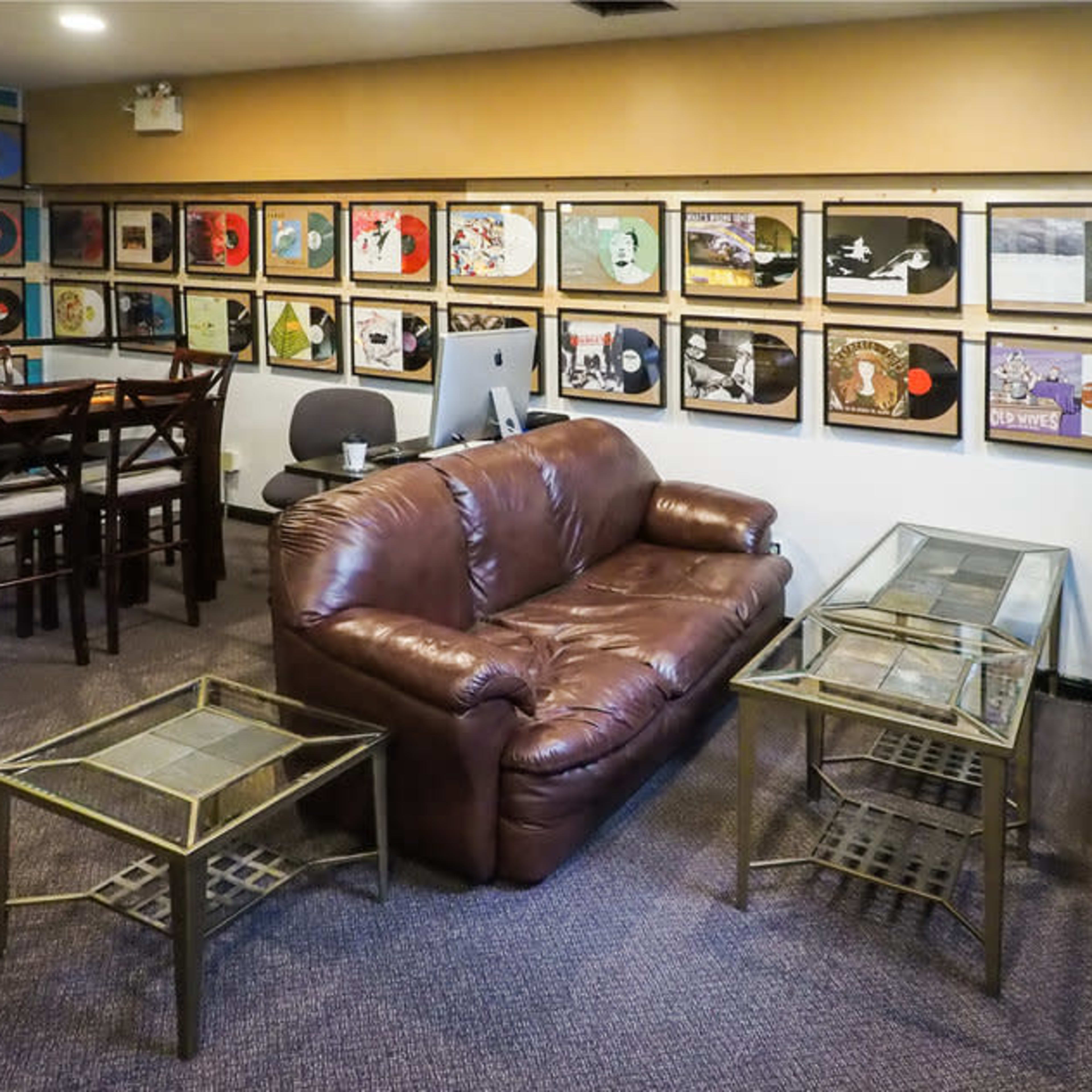 A room with a brown sofa and two glass-top tables, surrounded by framed album covers on the walls and a dining table in the corner.