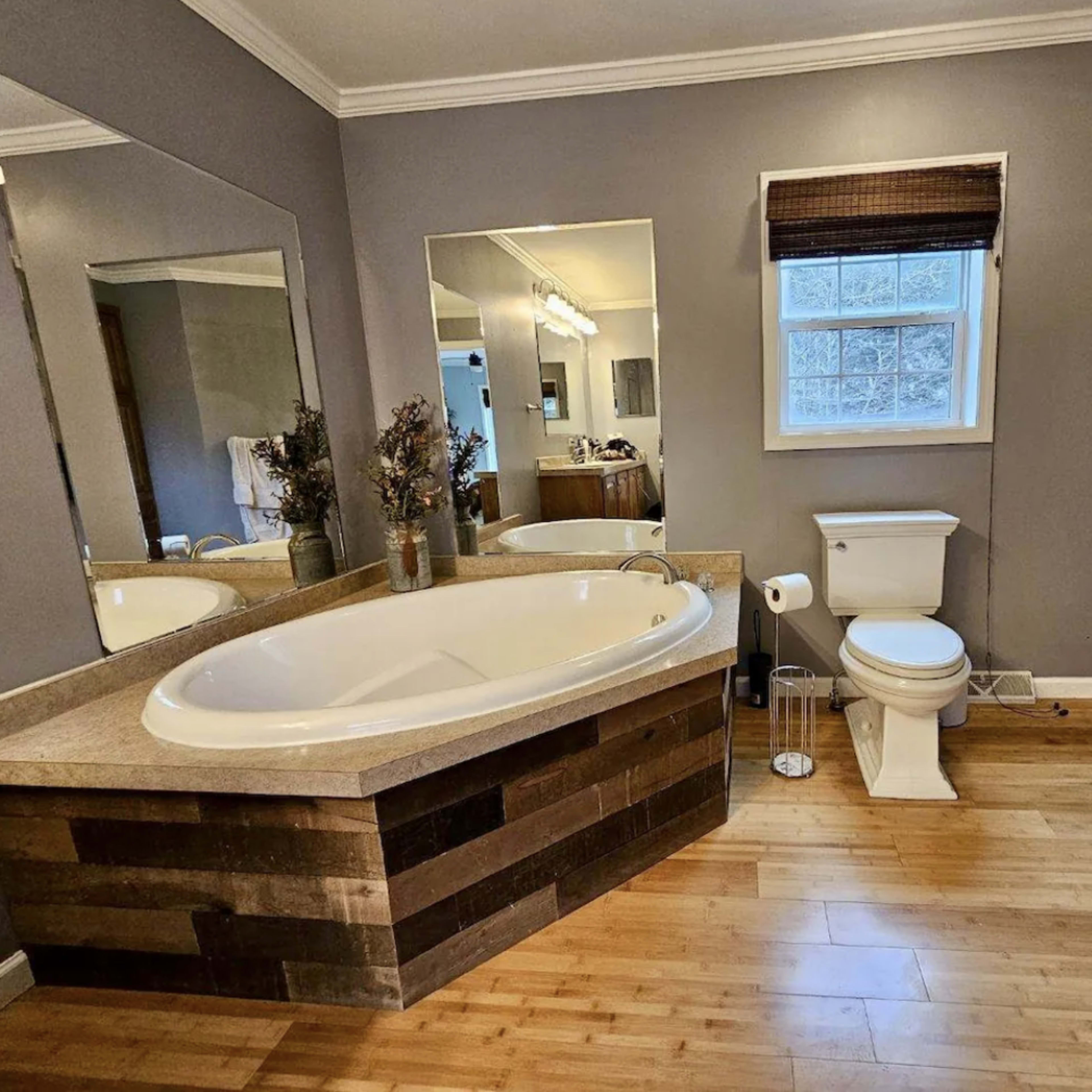 A bathroom featuring a large oval bathtub, a toilet, two mirrors, and a window with a bamboo blind.