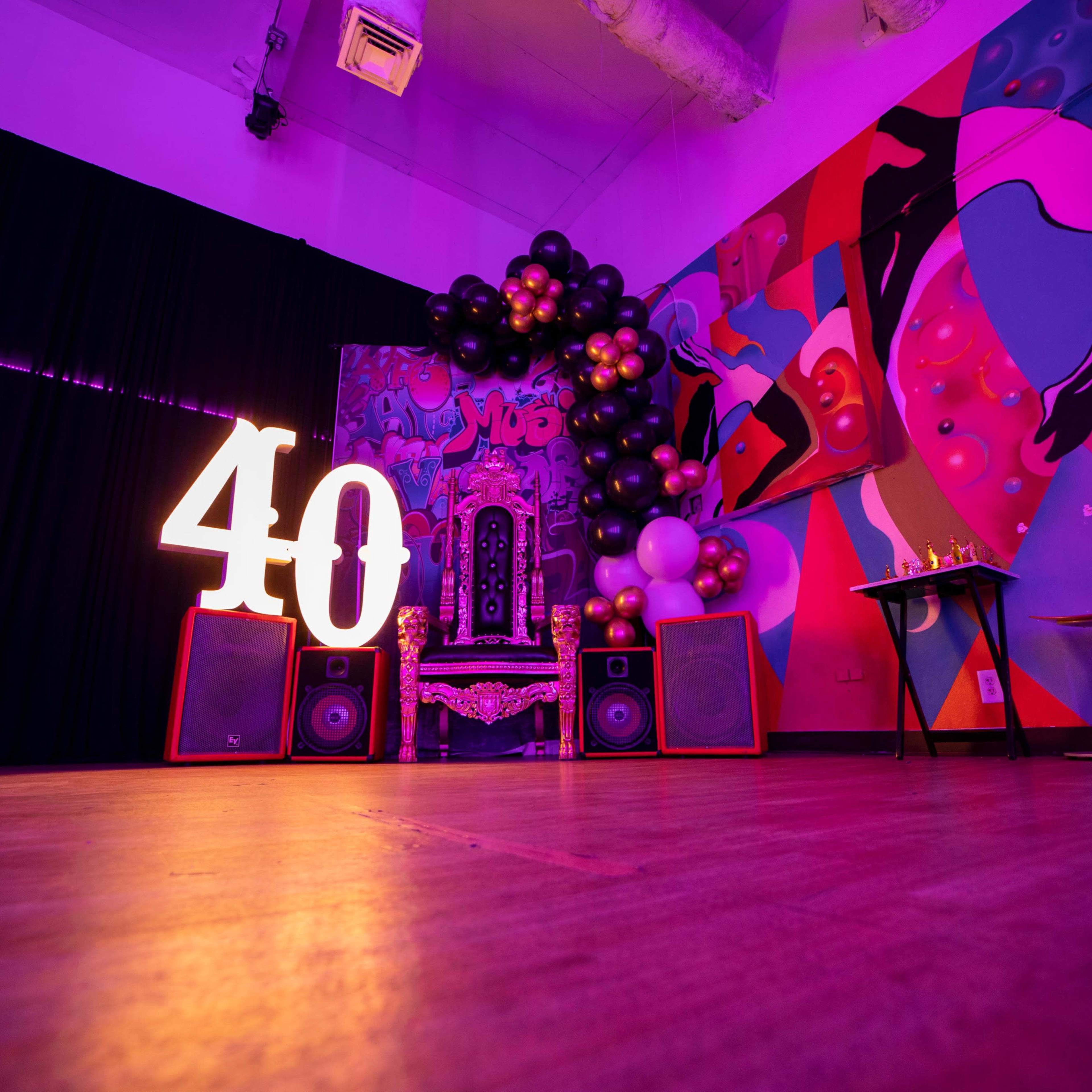 A party setting decorated with a large illuminated "40," a throne-like chair, speakers, and colorful balloons against a vibrant wall mural.