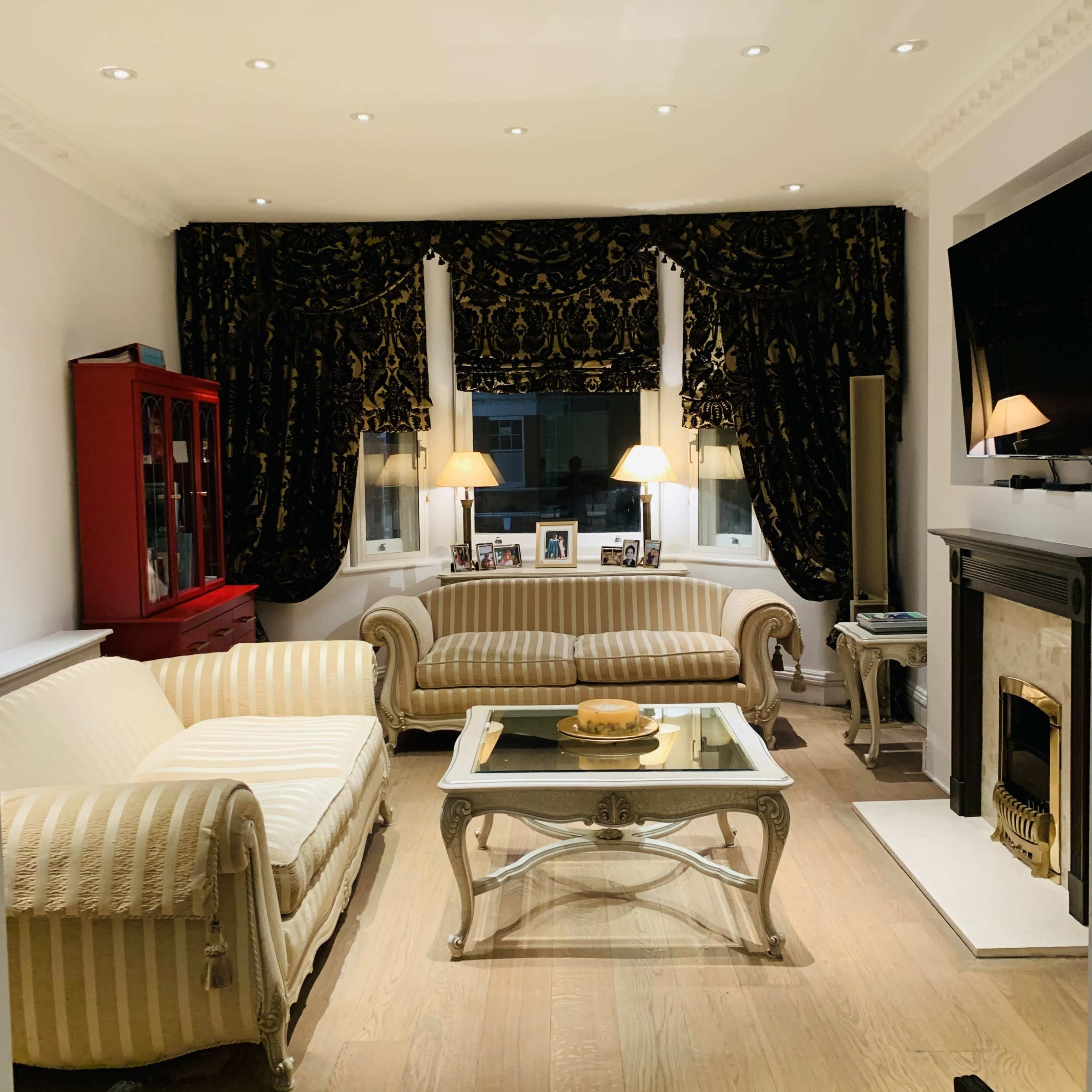 The living room features two striped sofas, a coffee table with a decorative centerpiece, and a fireplace, framed by dark curtains and illuminated by lamps.
