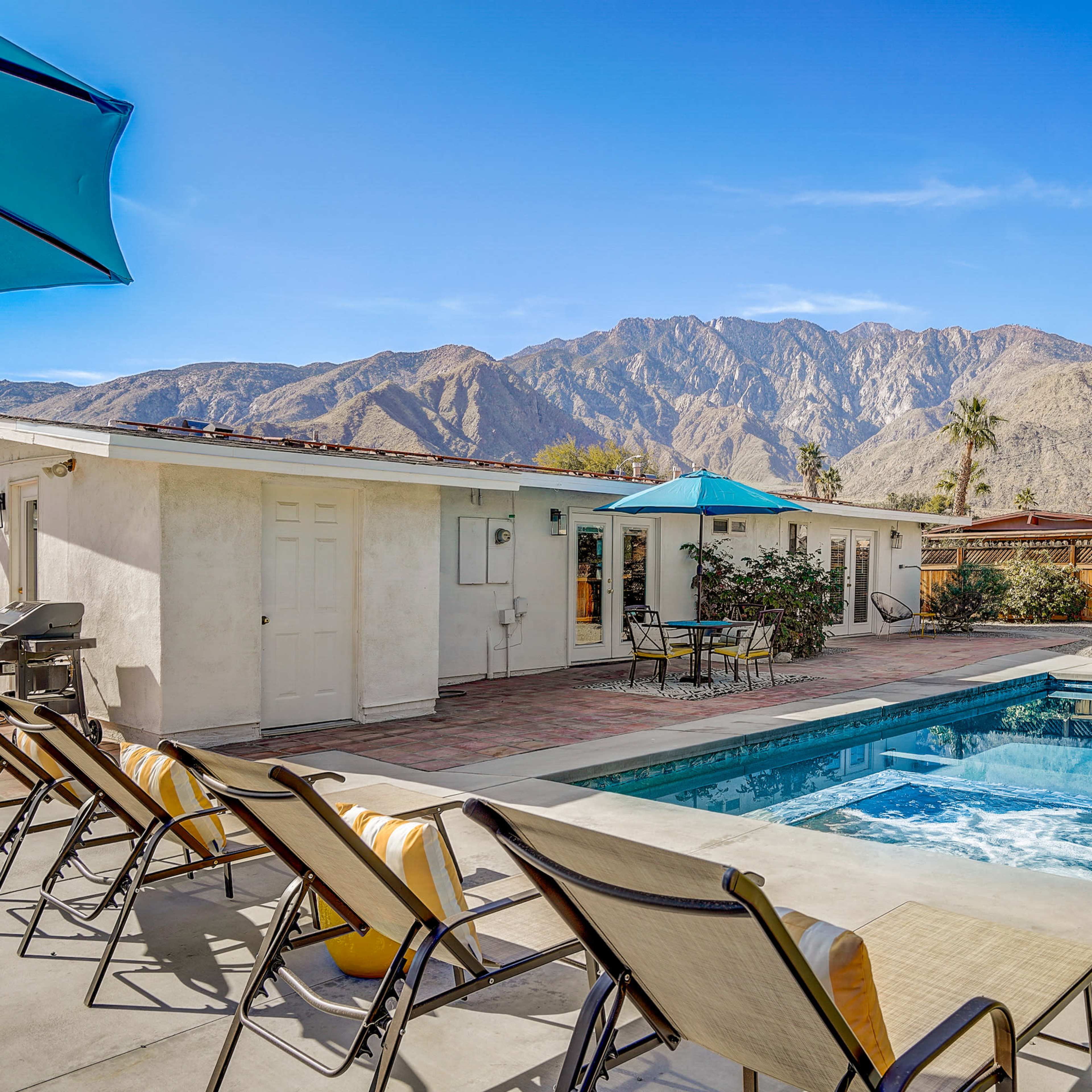 A shaded patio with lounge chairs surrounds a pool, with mountains in the background and palm trees lining the property.