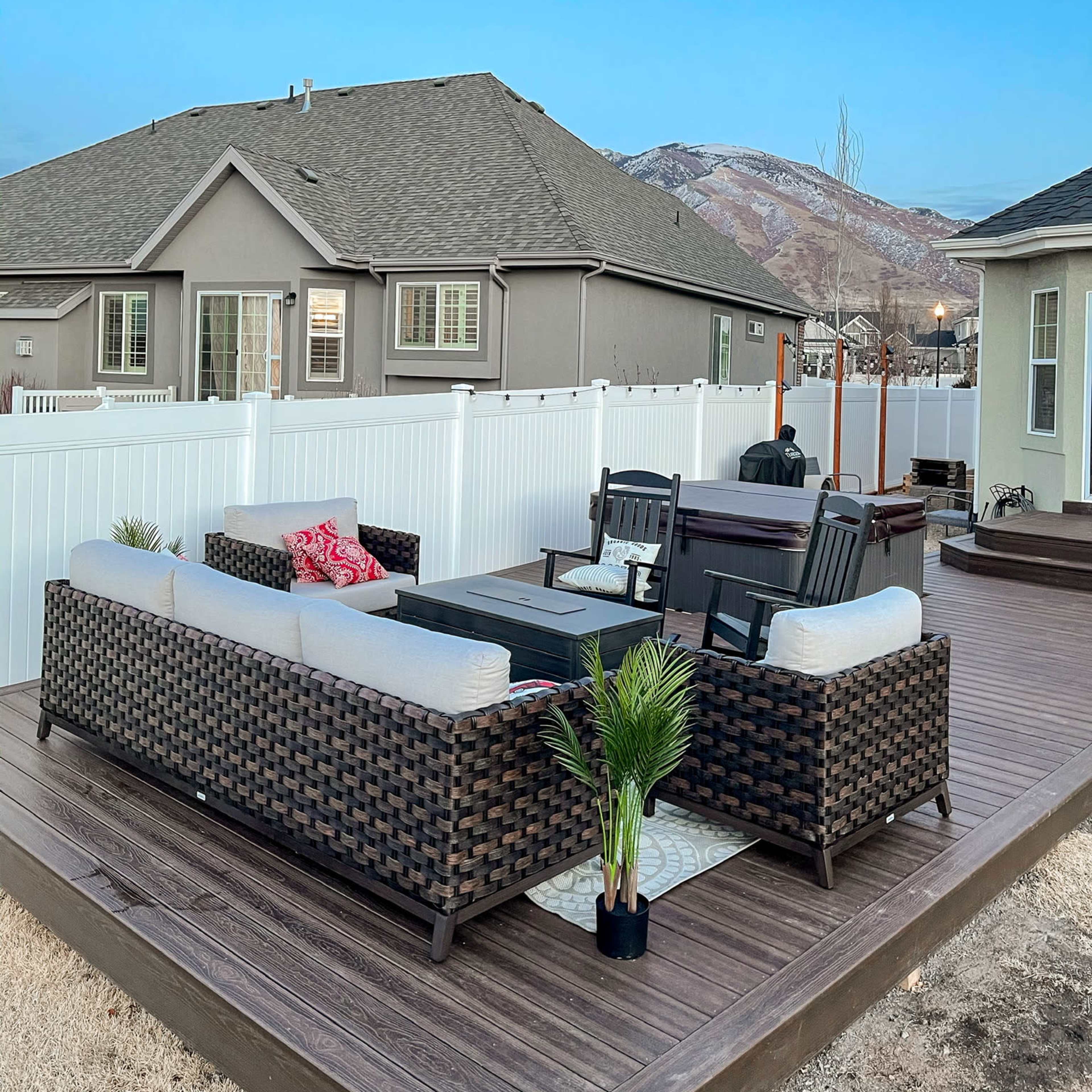 A patio area features a seating arrangement with wicker furniture and a small table, surrounded by a fenced yard and houses in the background.
