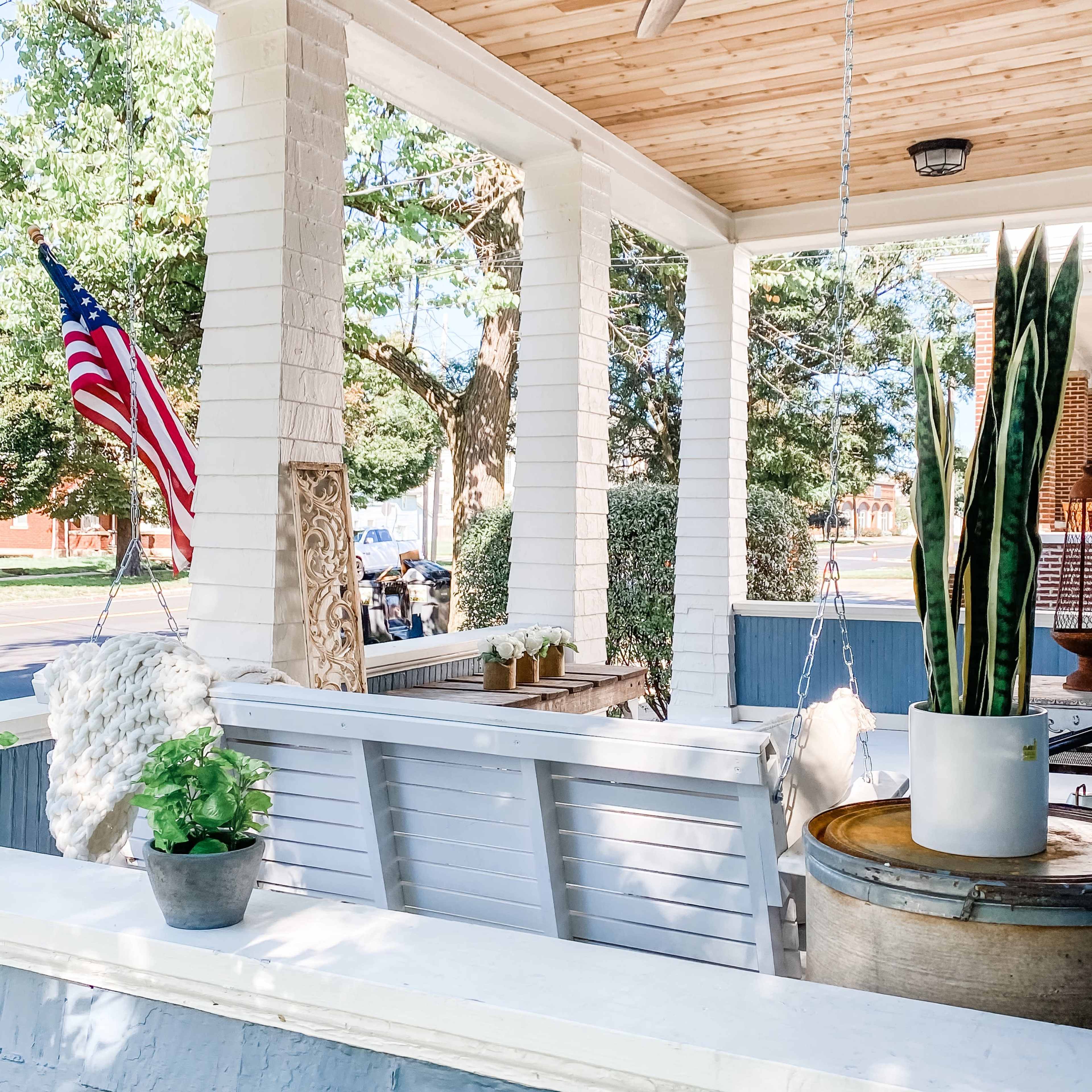 A porch with white brick columns, a ceiling fan, a hanging swing bench, an American flag, plants, and wooden accents.