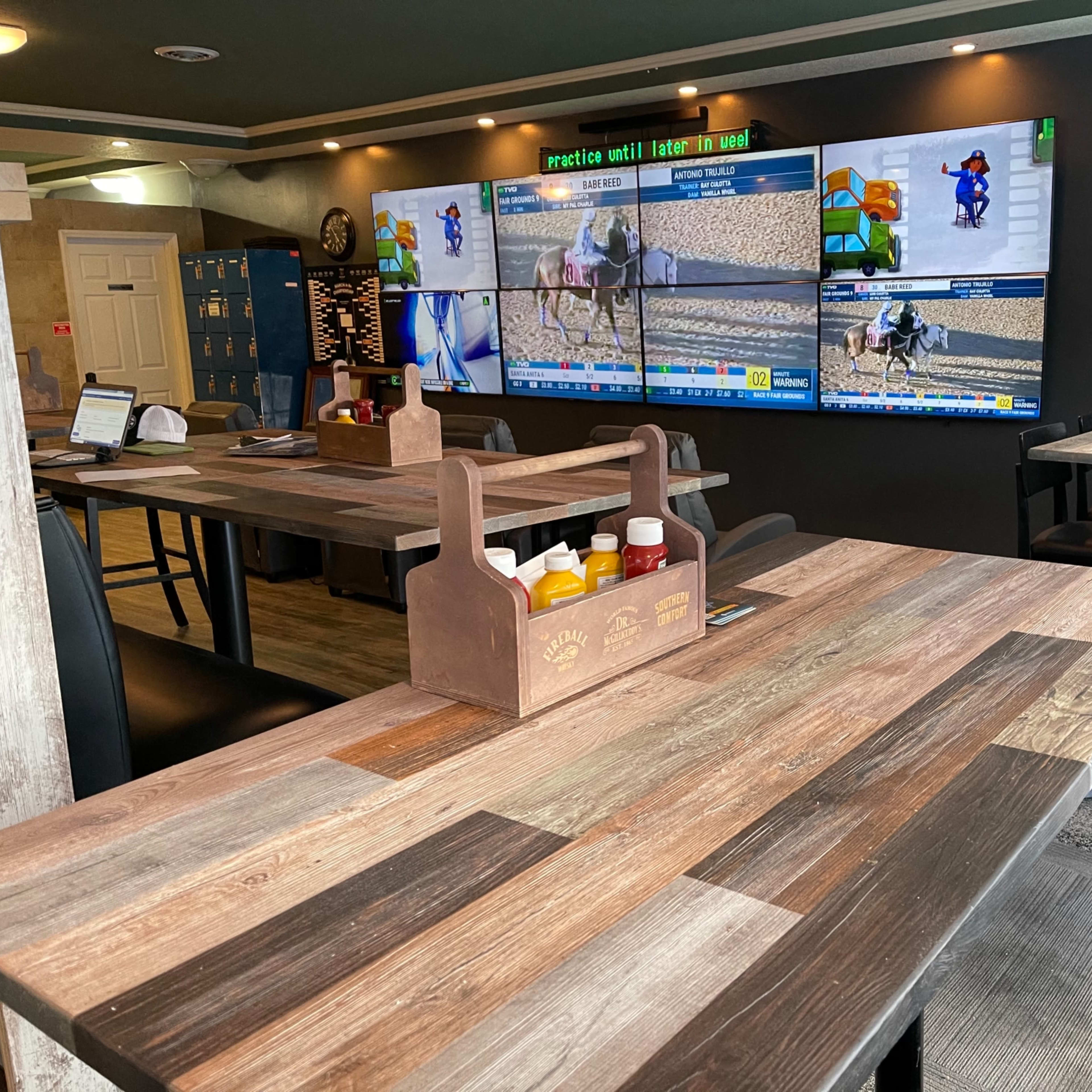A rustic dining area with wooden tables and a condiment carrier, featuring multiple screens displaying horse racing events on the wall.