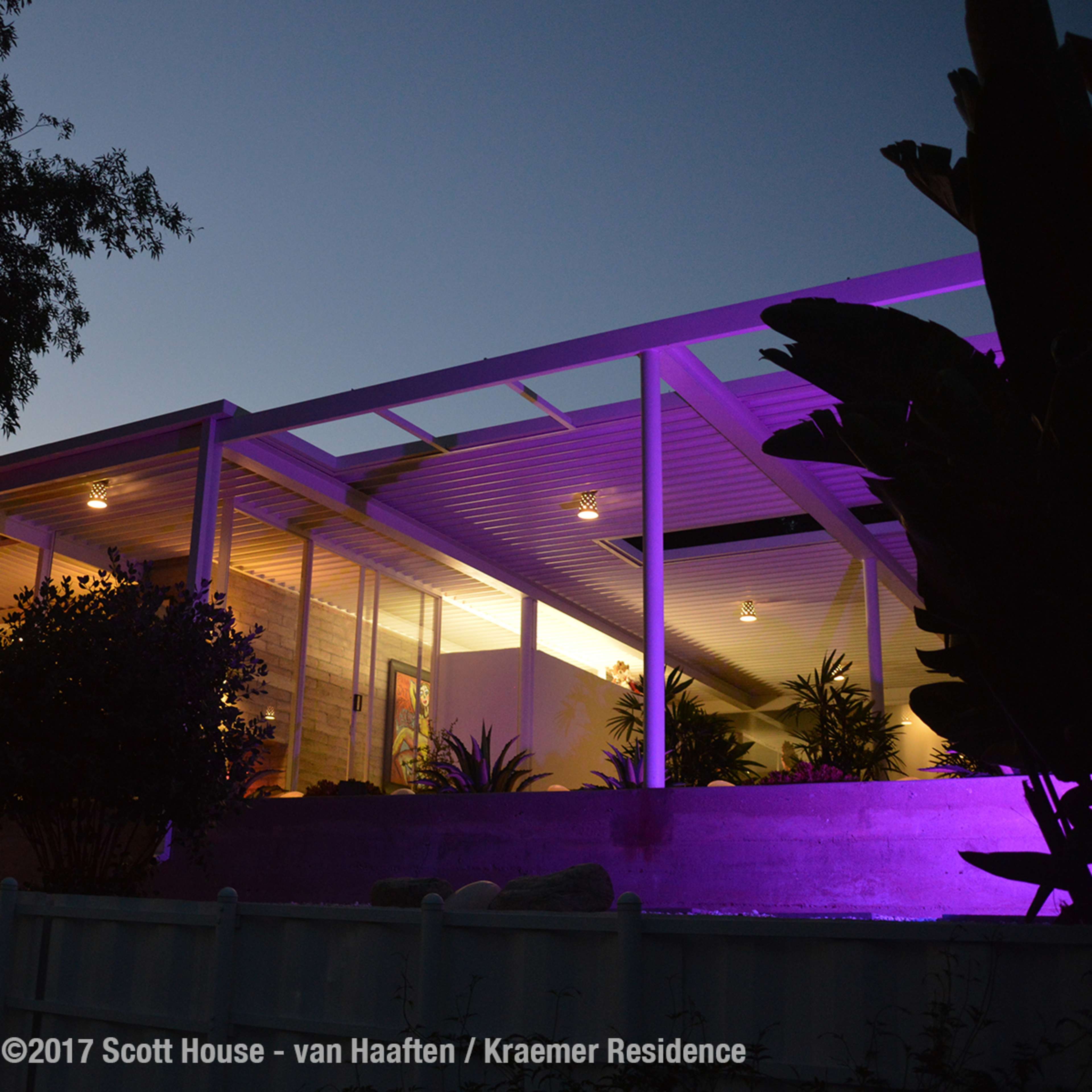A modern house with large glass walls, illuminated by purple lighting, amid surrounding trees and landscaping.