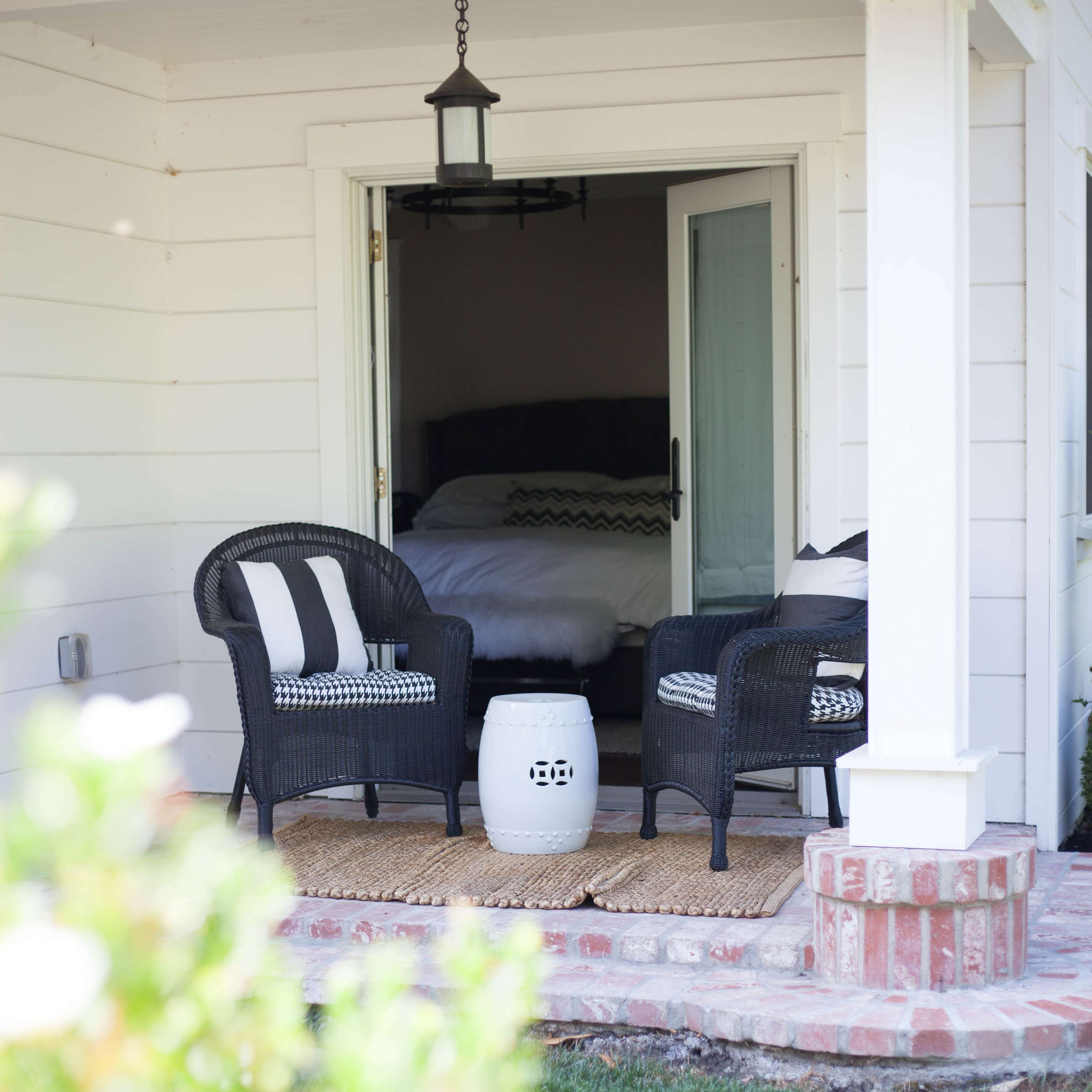 A porch with two black wicker chairs and a small round table is accessible through an open door leading into a bedroom.