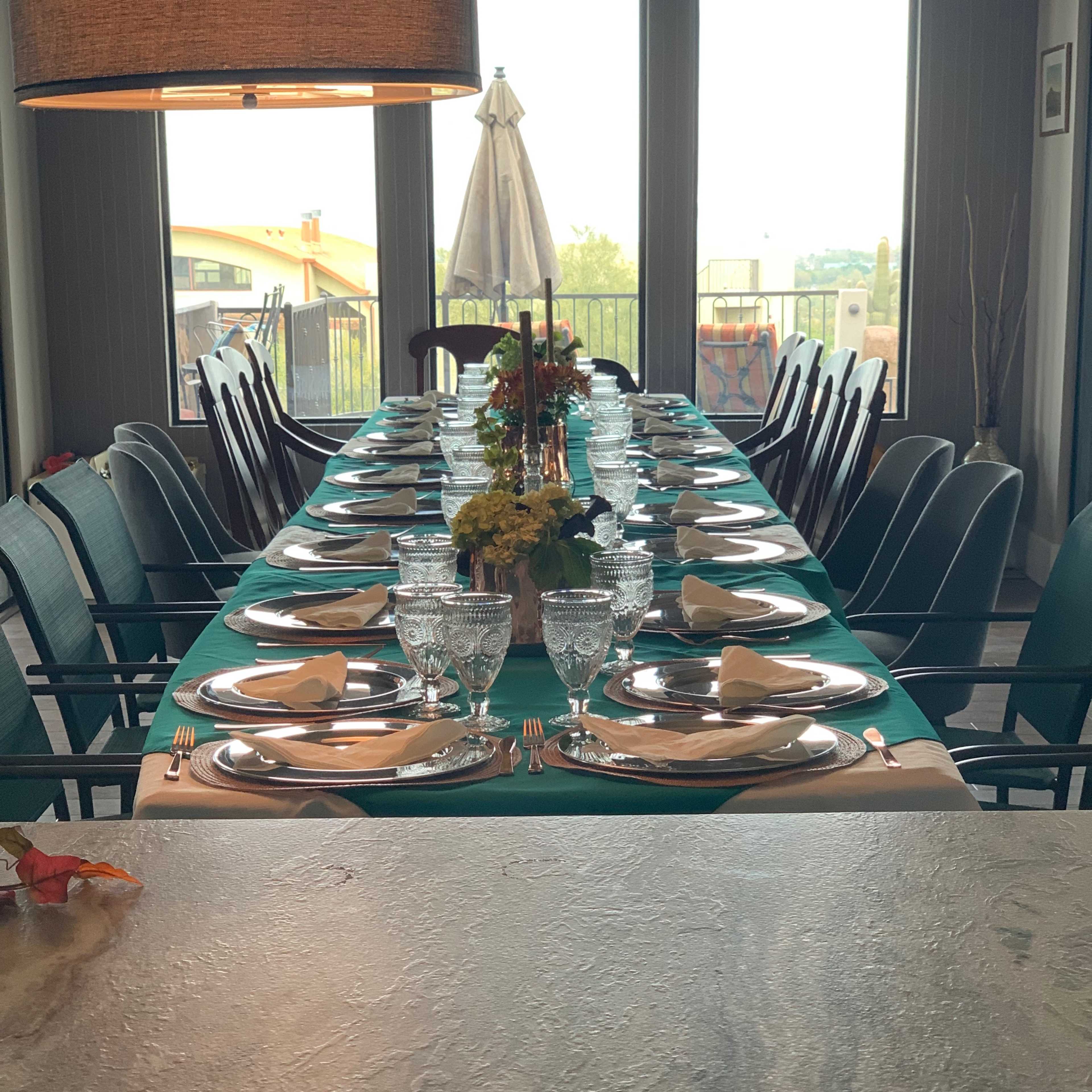 A long dining table is set with plates, glasses, and napkins, surrounded by chairs, under a large window with a view outside.