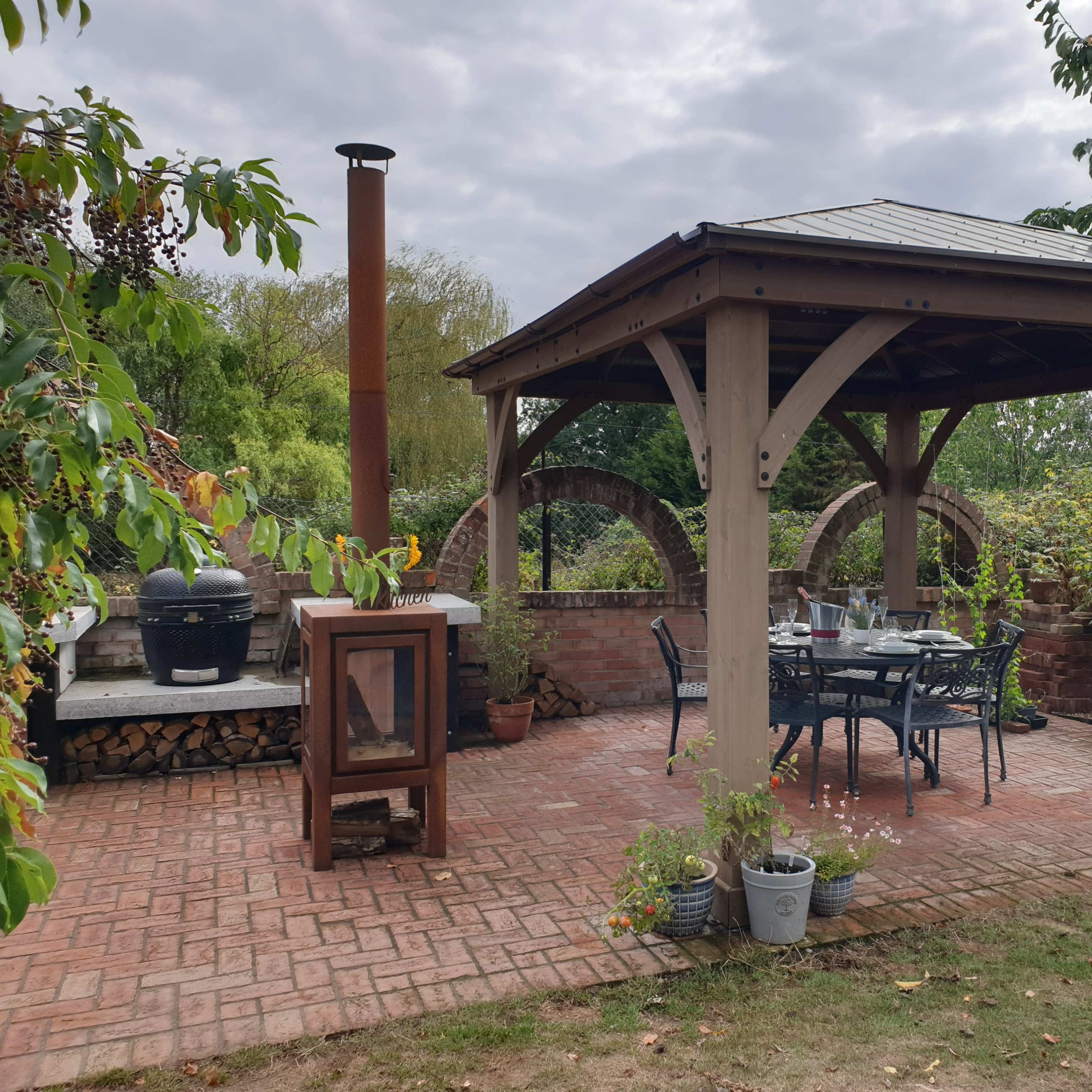 A wooden gazebo with a metal roof is surrounded by plants and features a dining set on a brick patio.