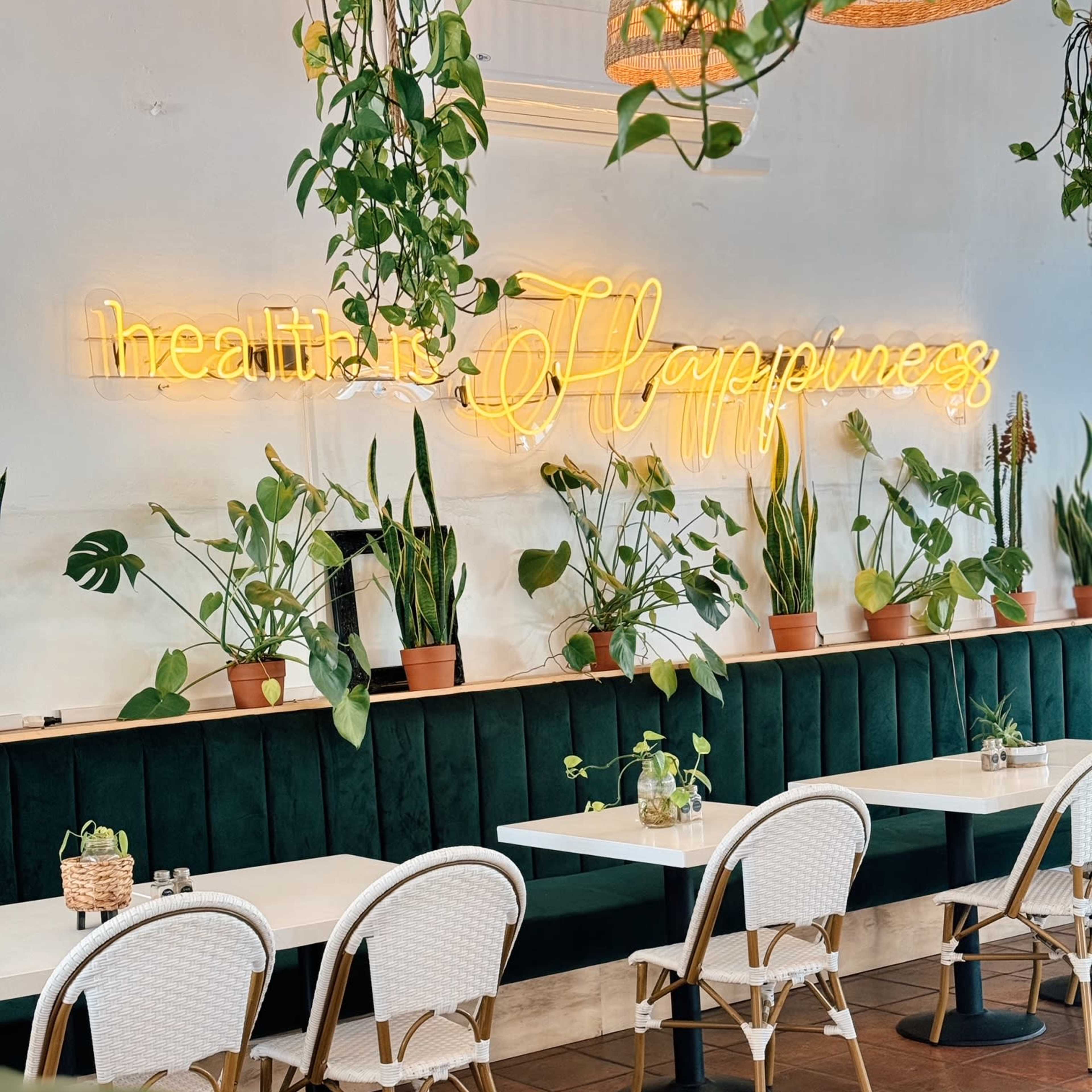 A stylish cafe interior with green velvet seating, light wooden tables, and potted plants, featuring a neon sign that reads "health is Happiness."