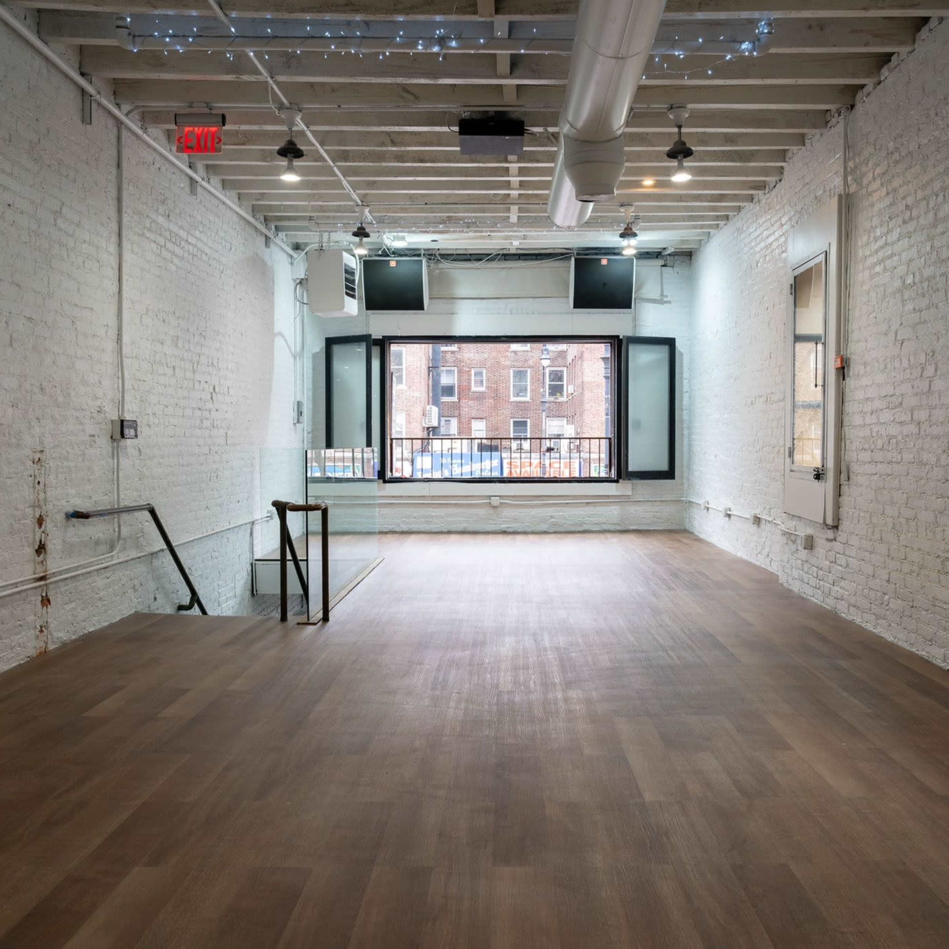 A spacious, empty room with white brick walls and hardwood flooring features large windows overlooking a street.