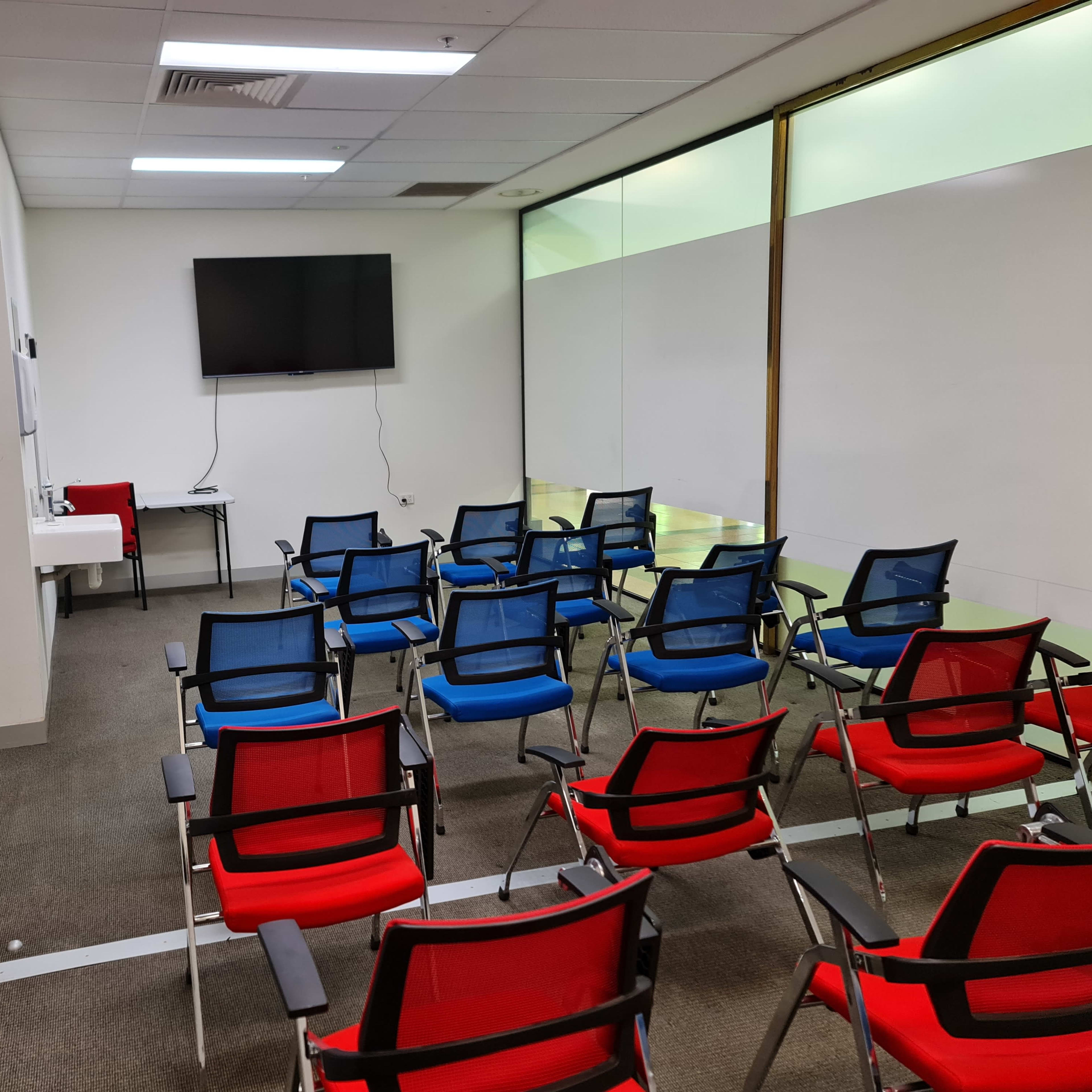 A meeting room with red and blue chairs arranged in rows facing a wall-mounted television and a table.