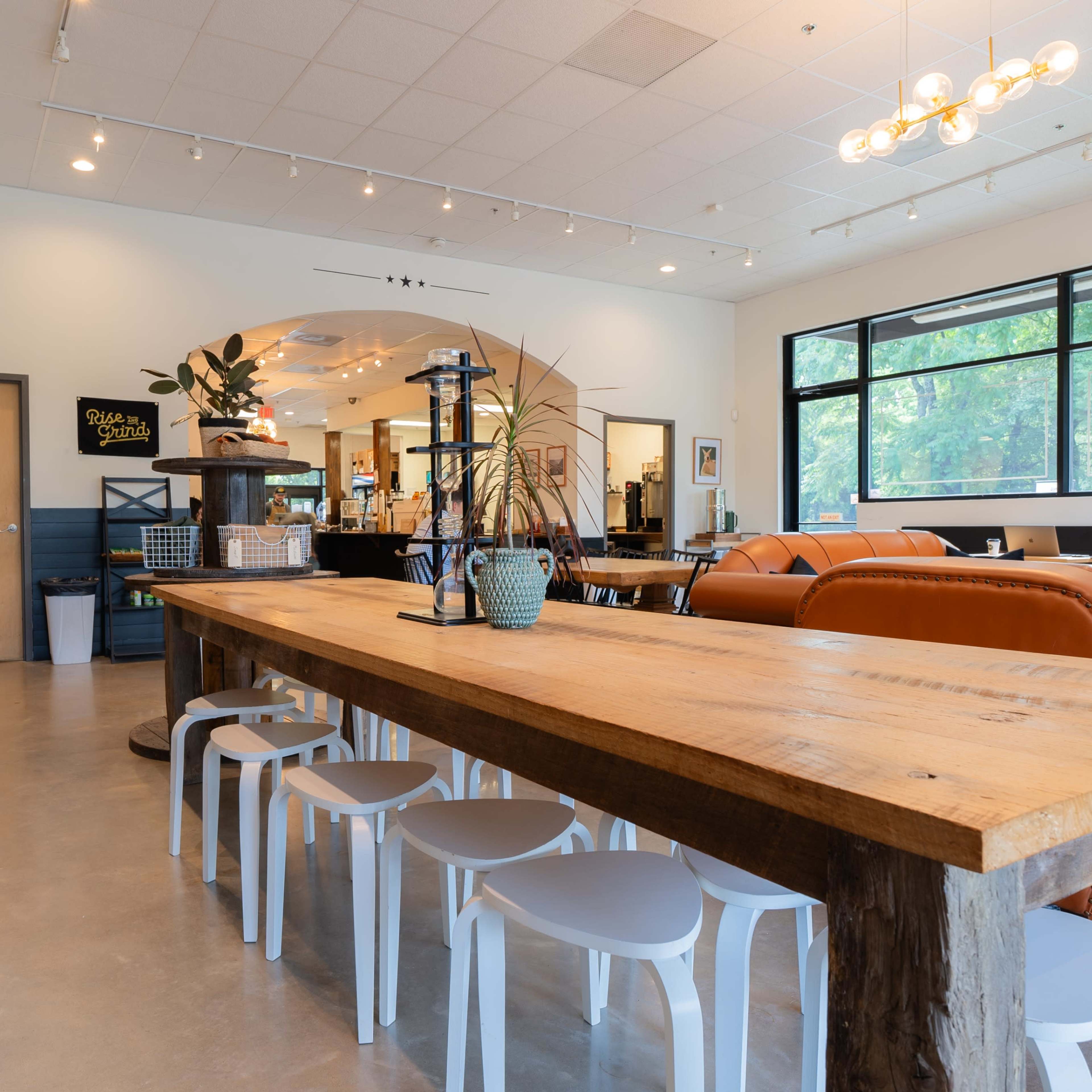 A modern café interior with a large wooden communal table surrounded by white chairs, orange seating areas, and large windows letting in natural light.