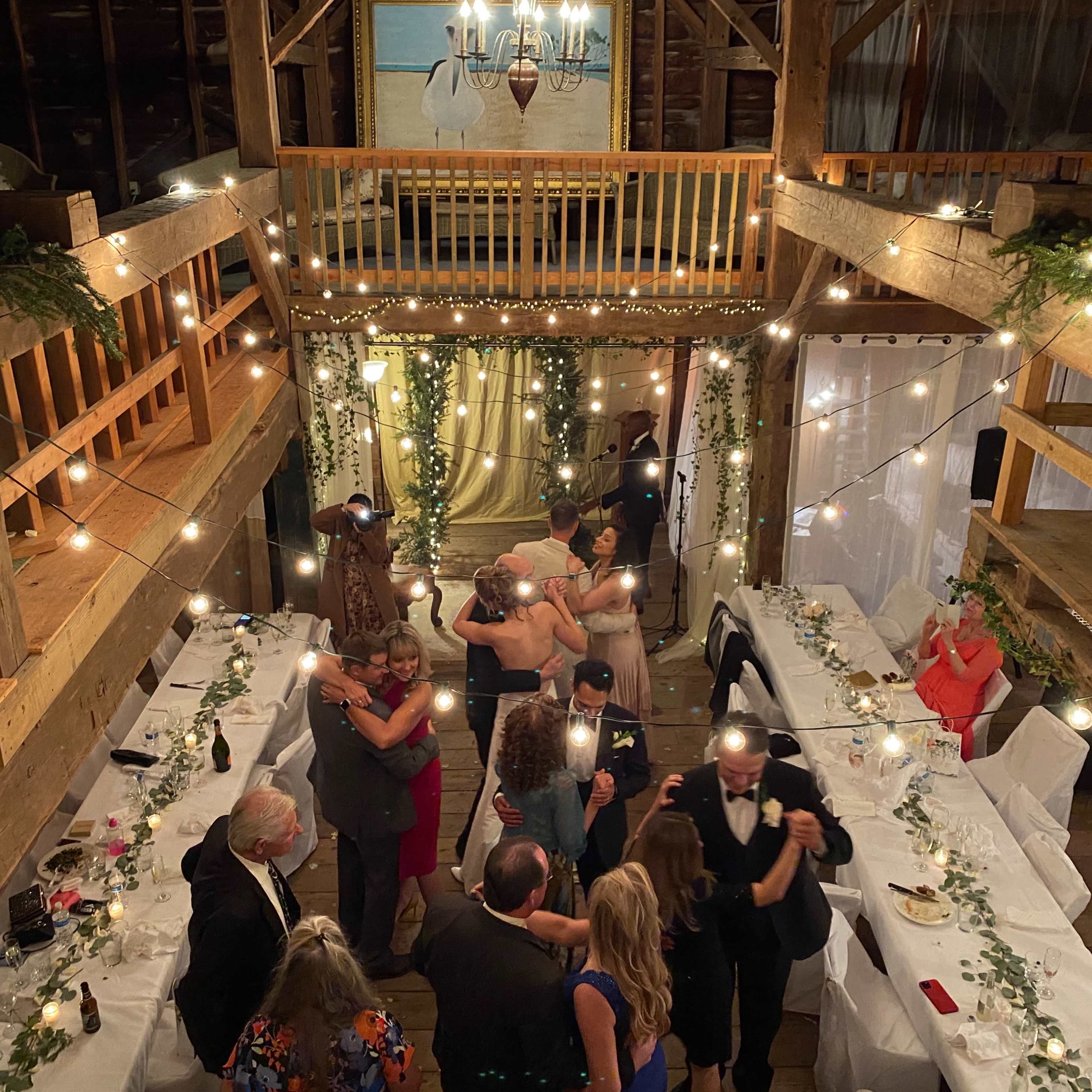 A large group of people dances under string lights in a barn decorated for a celebration, surrounded by tables set for a dinner.