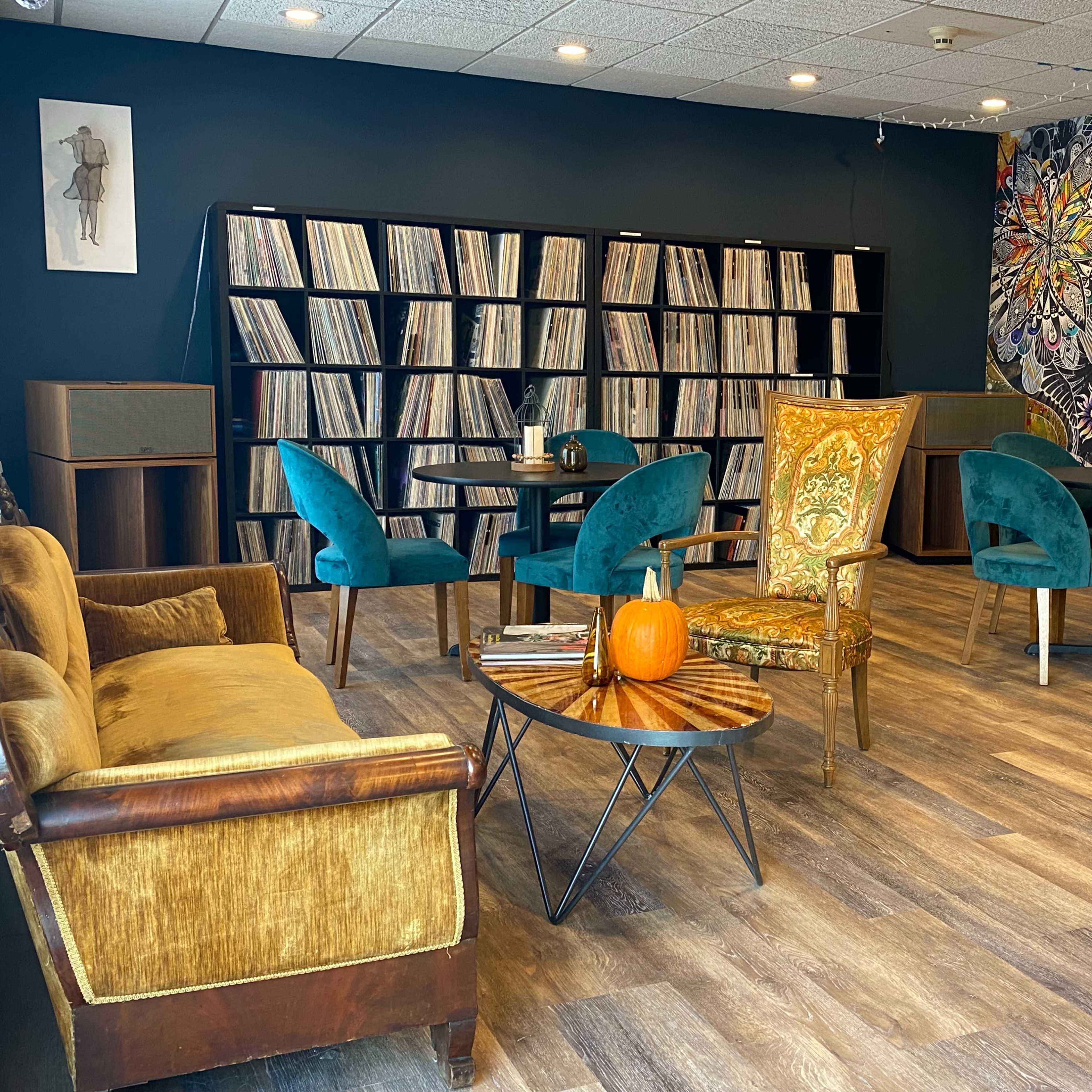 The room features a combination of vintage furniture, including a gold-accented chair and a brown couch, surrounded by shelves filled with vinyl records.