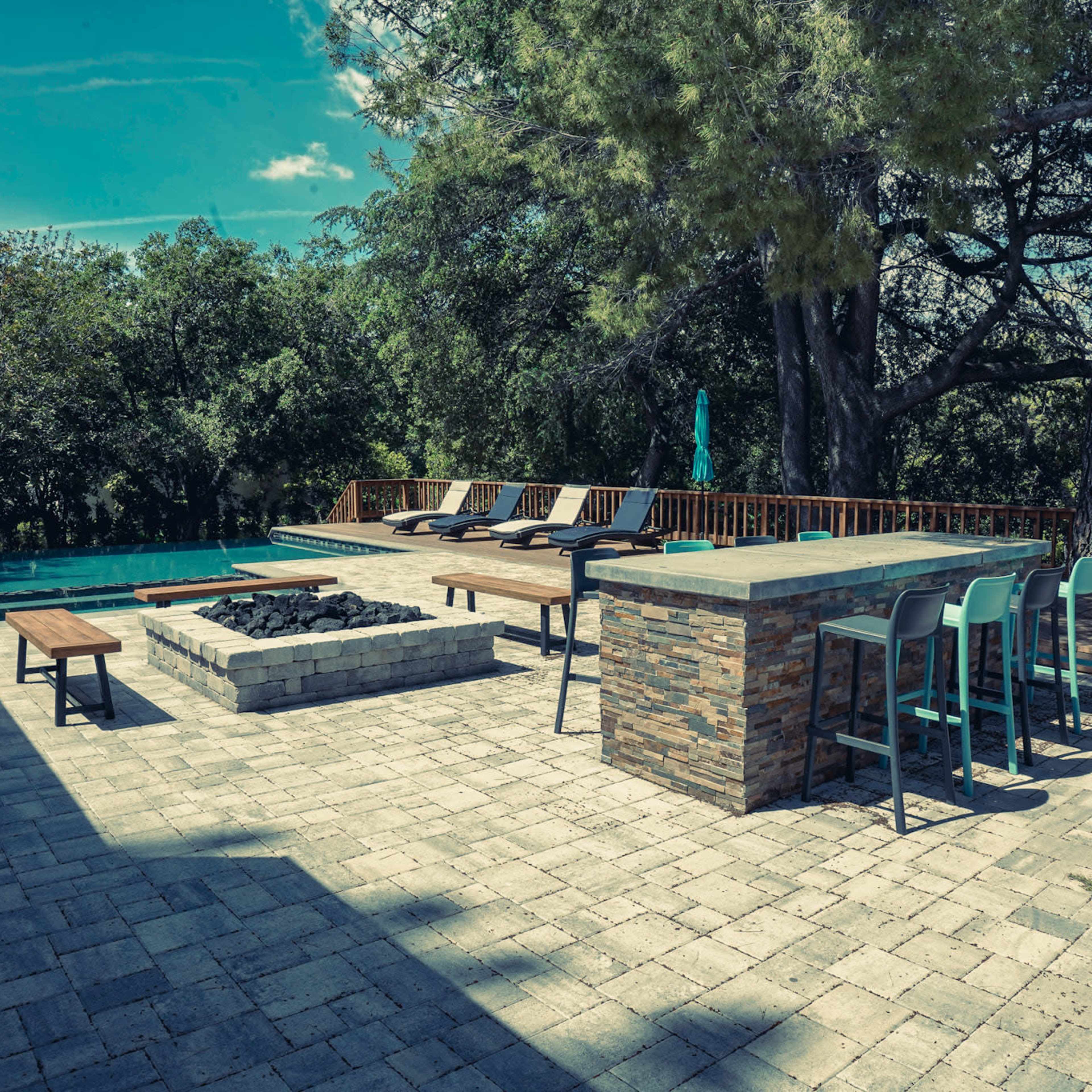 A spacious outdoor area featuring a swimming pool, lounge chairs, a fire pit, and a bar with high stools, surrounded by trees.