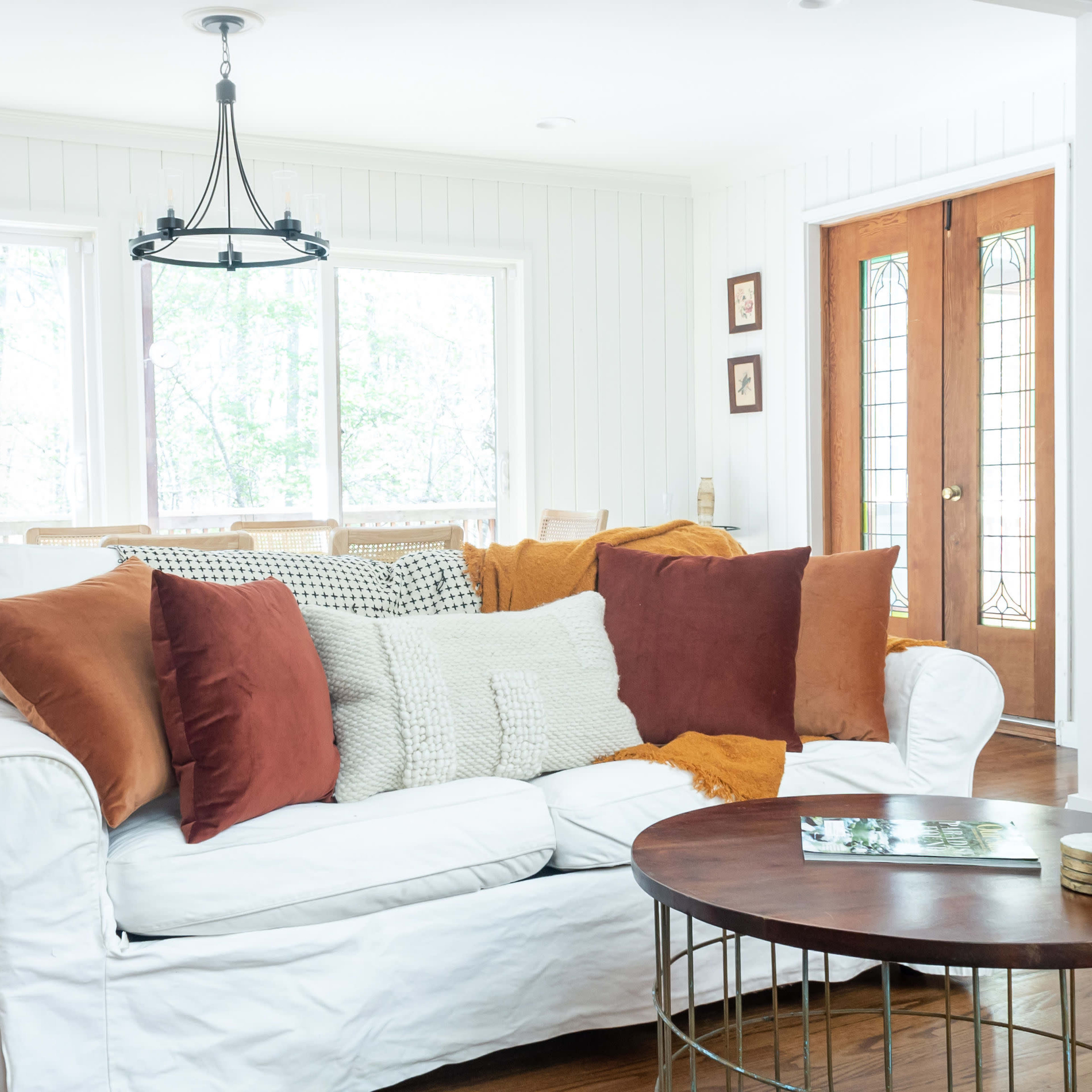 A white couch adorned with various textured pillows sits in a bright room with large windows and wooden doors.