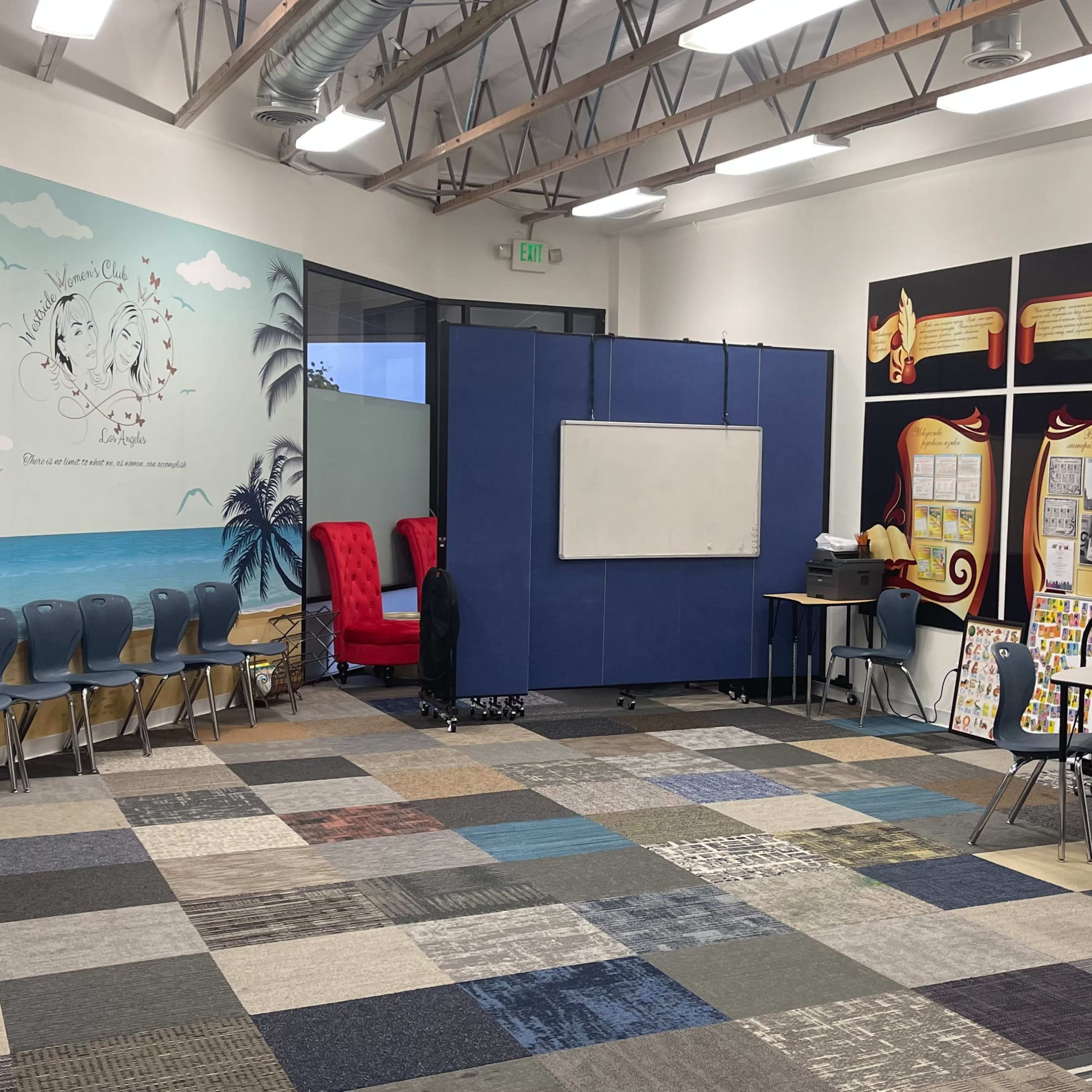 A classroom with blue partitions, colorful carpet tiles, and seating arrangements, including two red chairs and a table with various educational materials.
