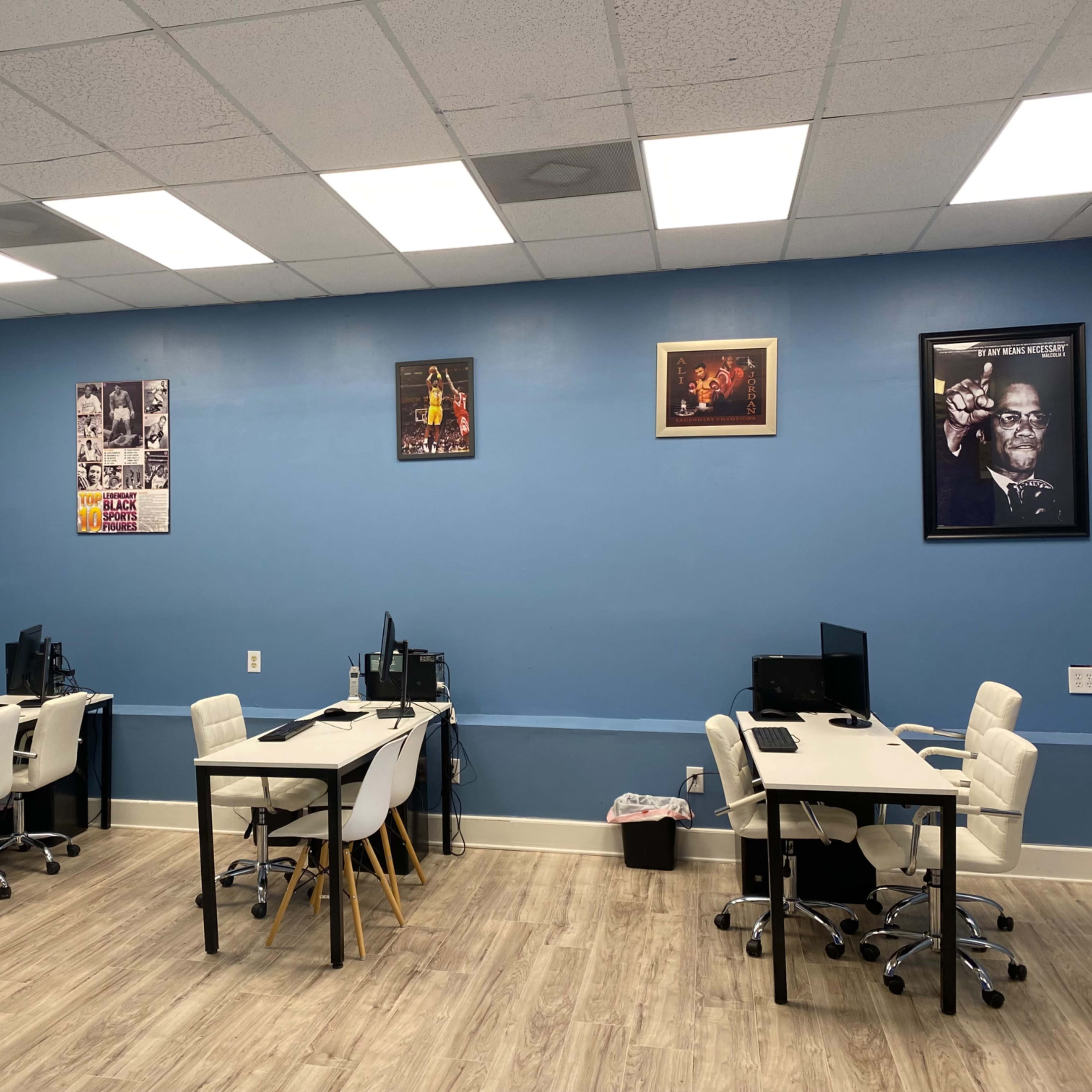 An office space with several white desks and chairs arranged in a blue-walled room, featuring posters on the walls and a television mounted on the left side.
