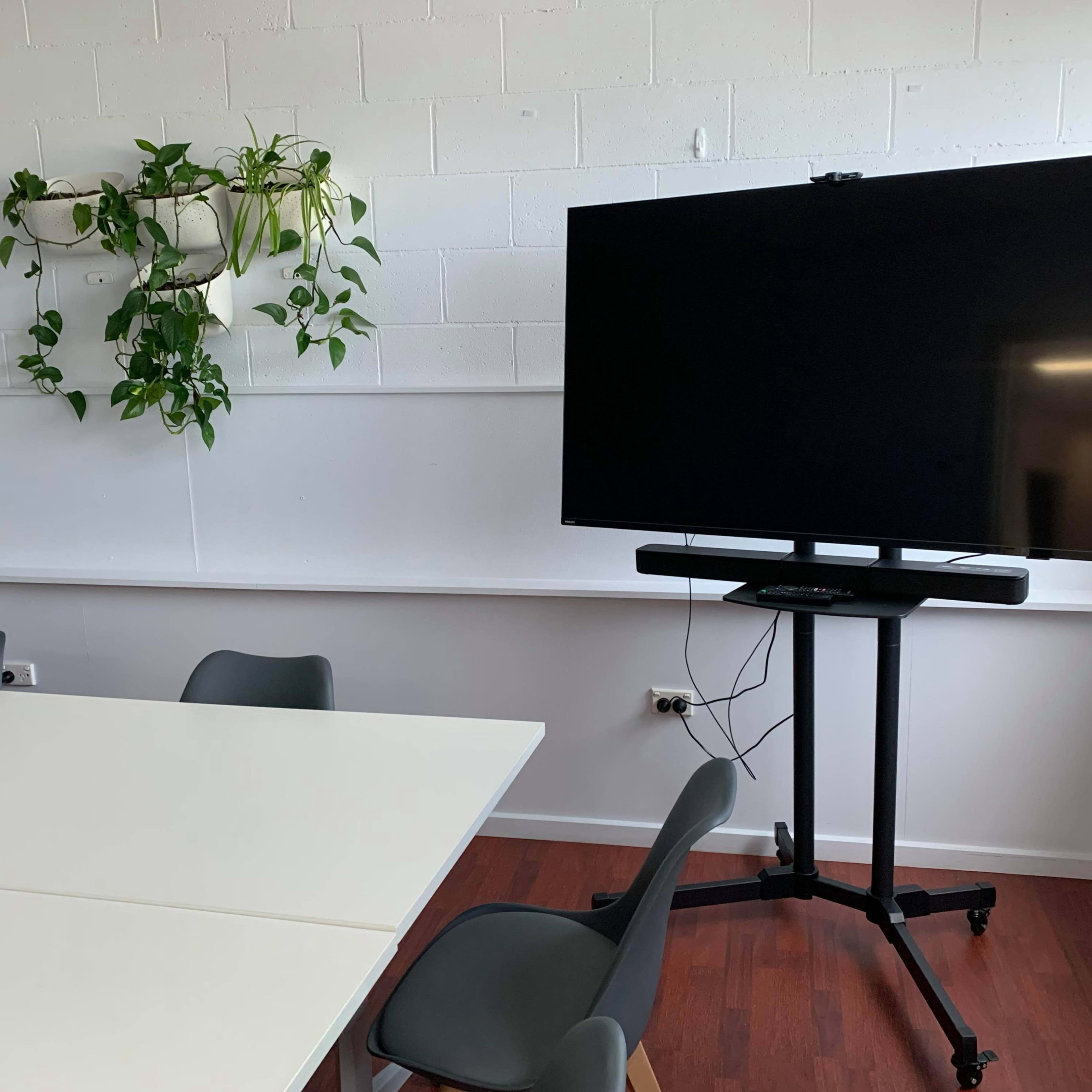 A modern meeting room features a large flat-screen television on a movable stand next to a table and green plants on the wall.