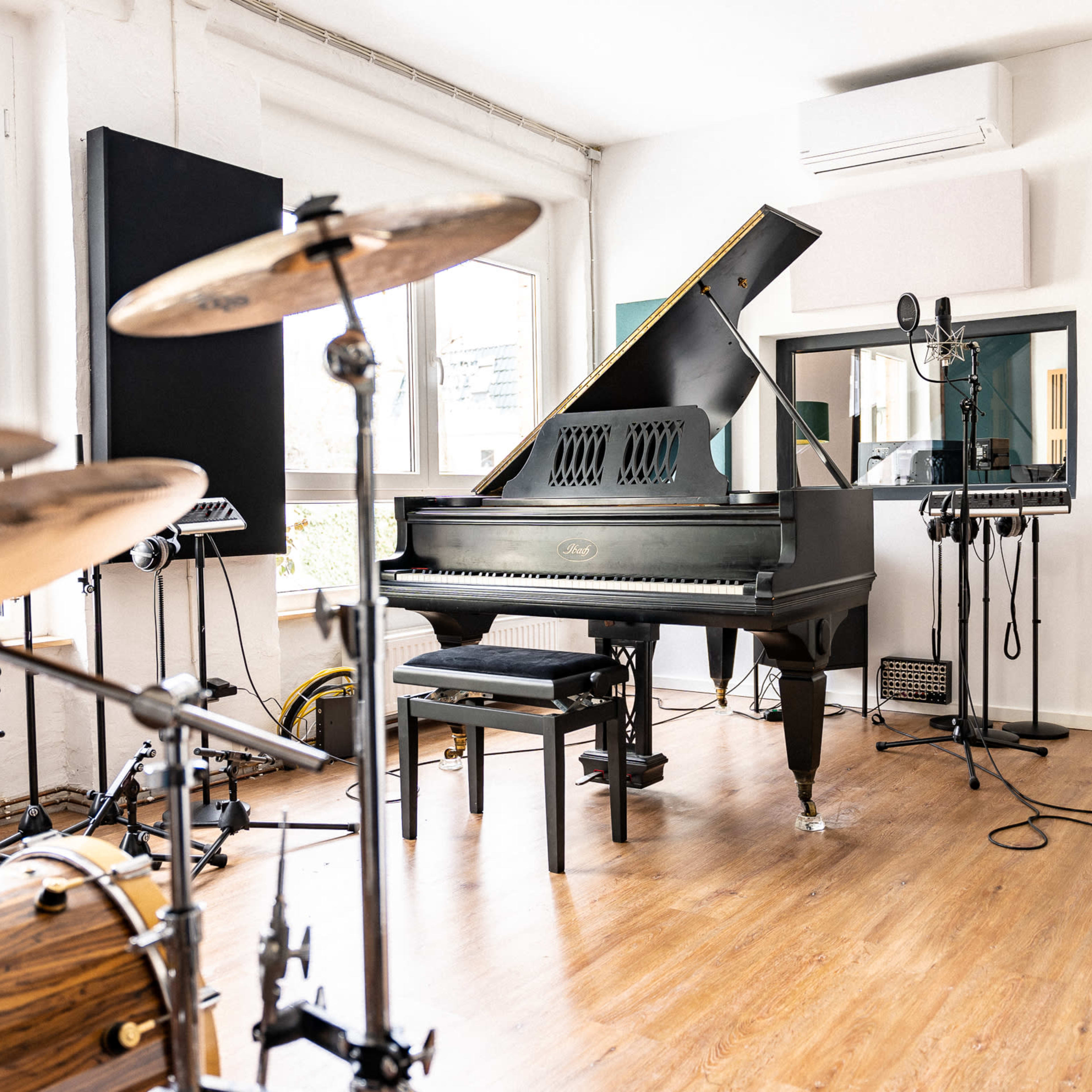 A music studio featuring a grand piano, a drum set, microphones, and sound equipment arranged in a bright room with large windows.