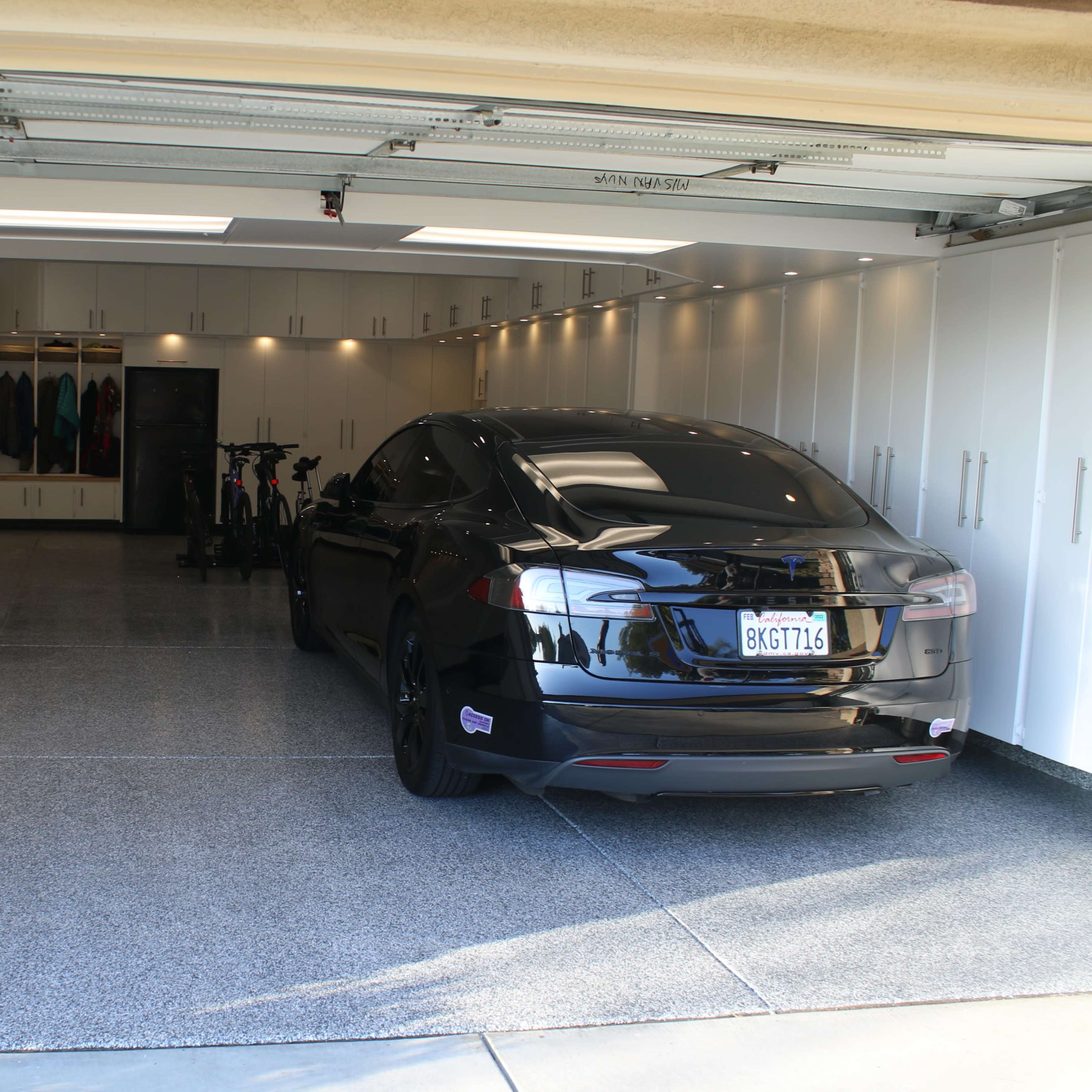 A black car is parked inside a spacious, organized garage featuring white cabinets and two bicycles on the side.