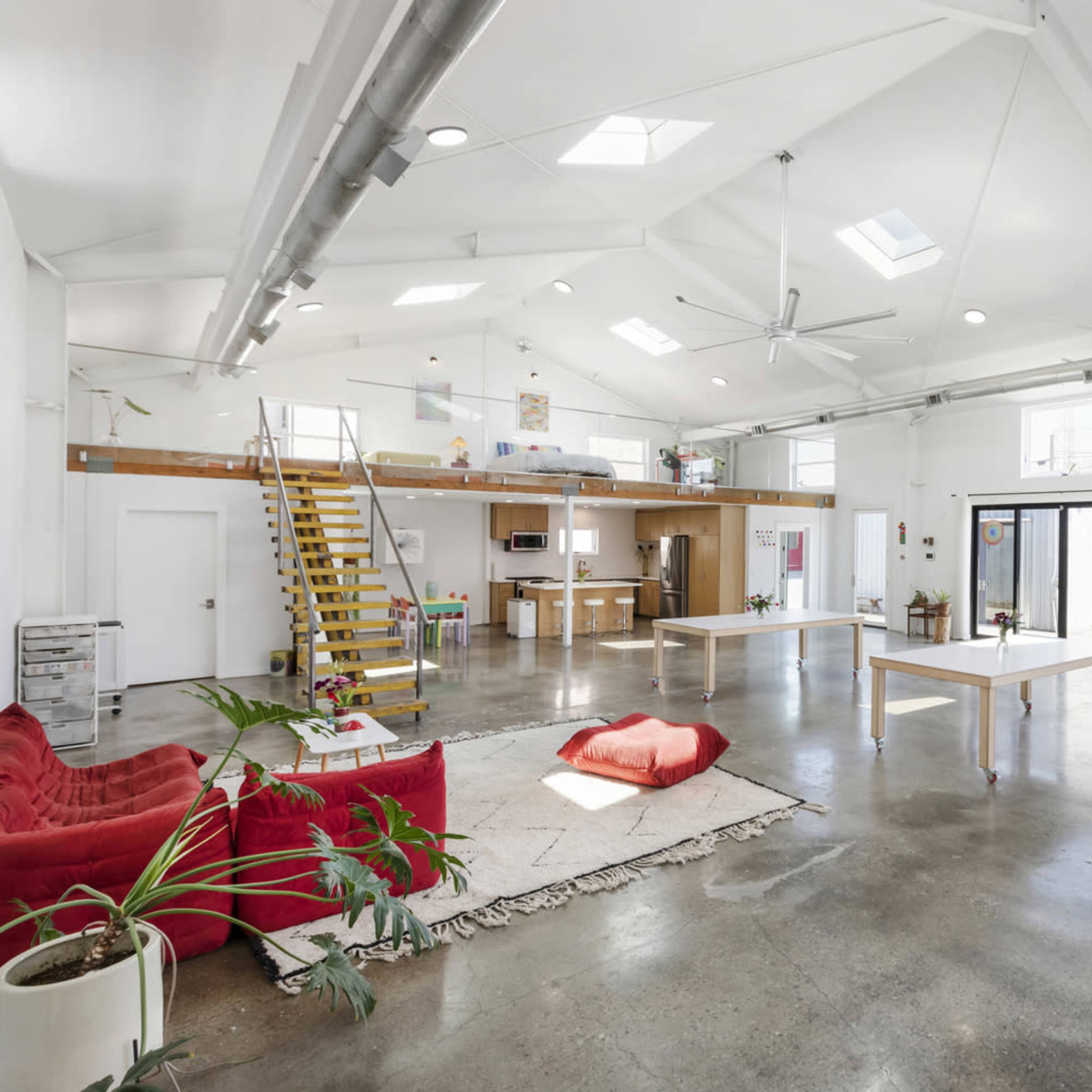 A spacious, well-lit interior of a modern studio with high ceilings, large windows, and minimalist furniture, including a red couch and white tables.