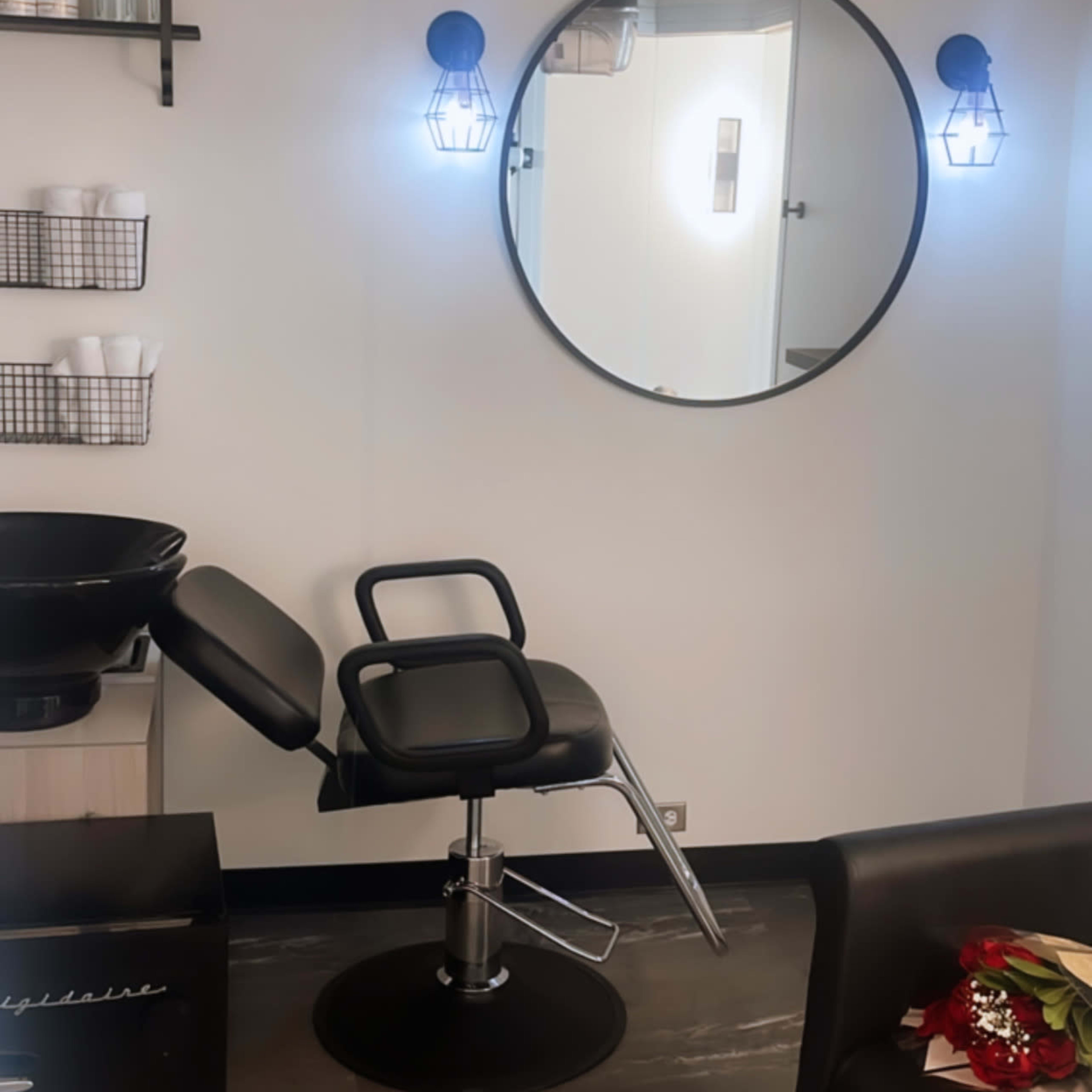 A modern hair salon interior with a black salon chair, a circular mirror, and wall-mounted shelves displaying various products.
