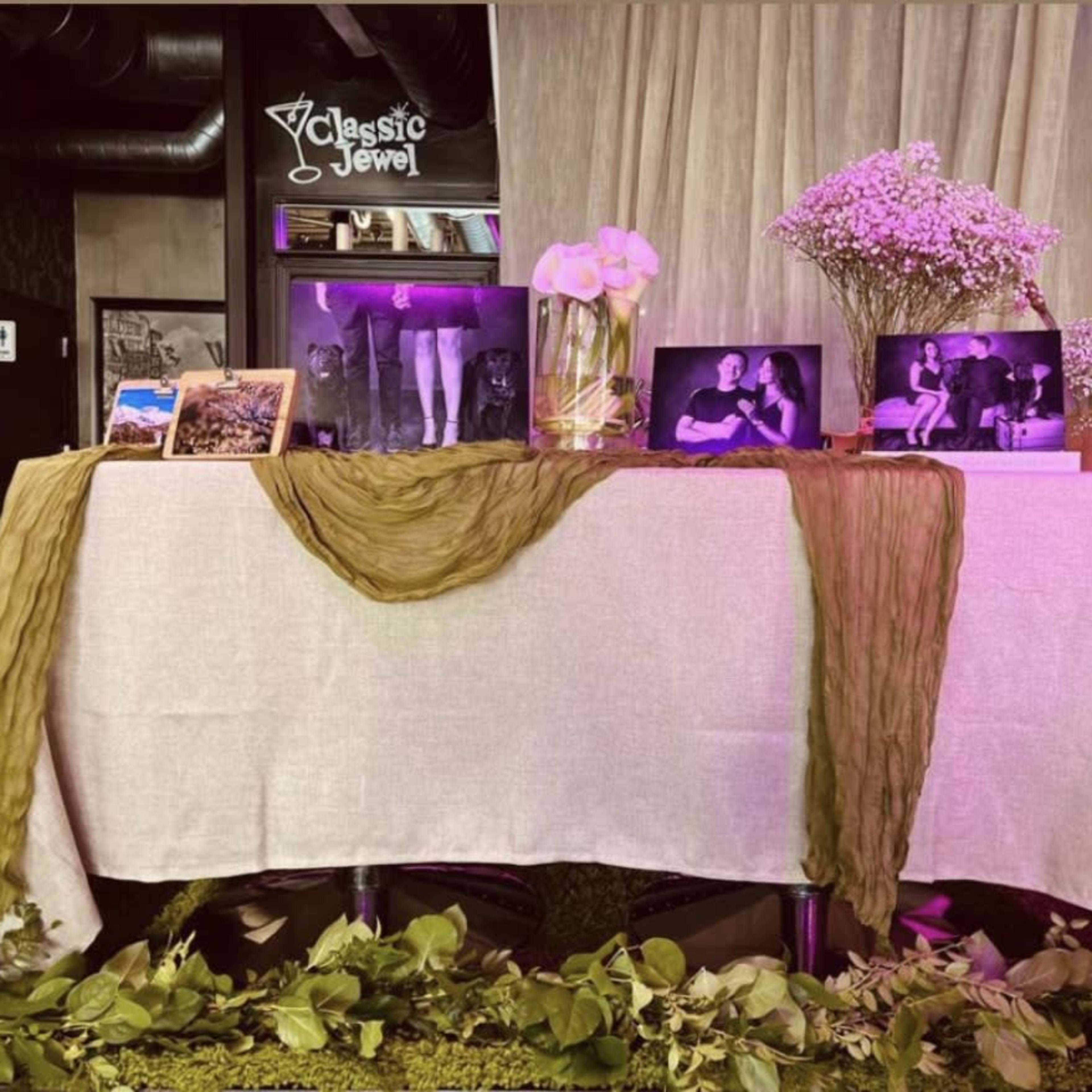 A table draped with a white cloth, adorned with photos and decorative items, set against a backdrop of soft lighting and fabric.