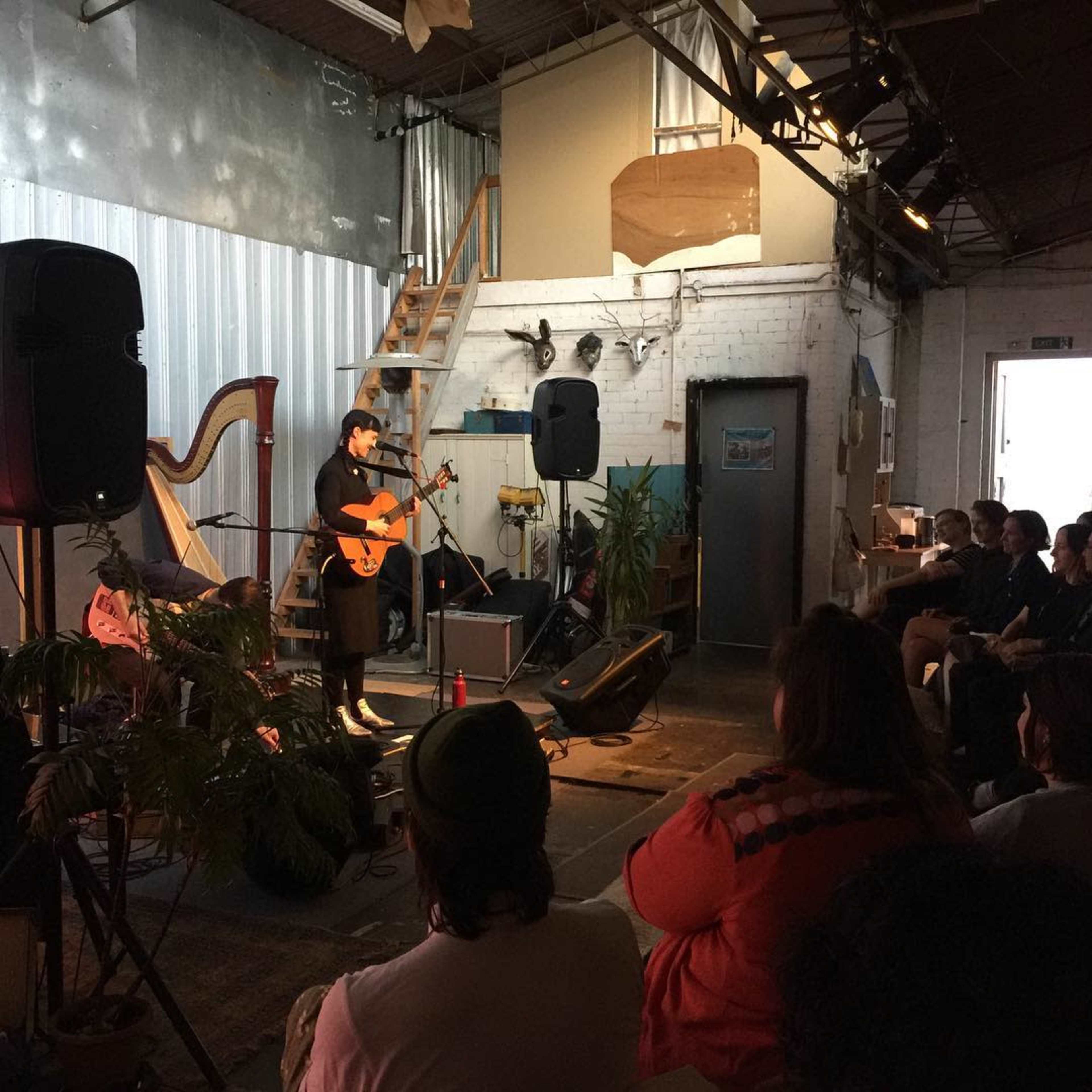 A musician performs on stage in a dimly lit warehouse setting with an audience seated in front.