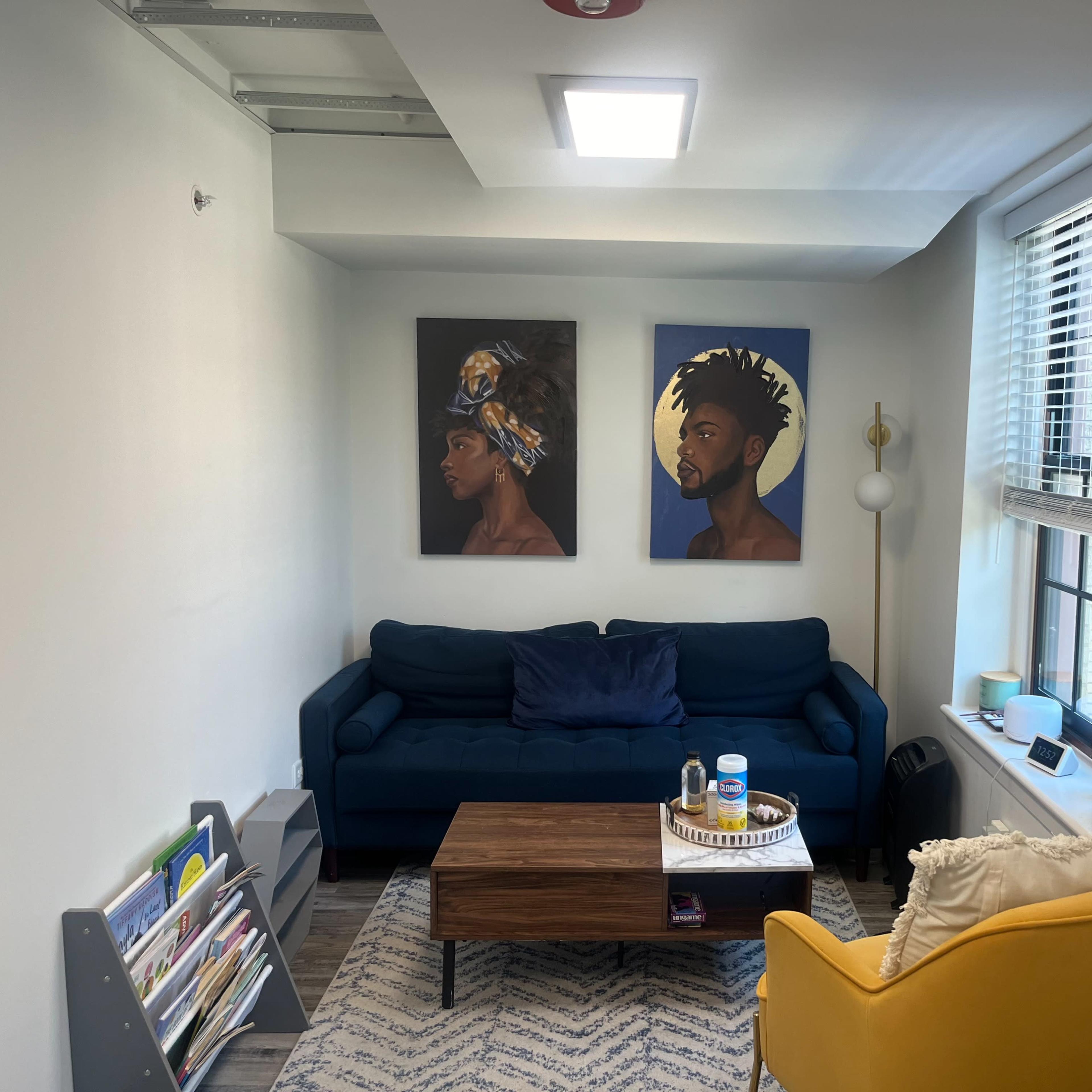 A small living space featuring a blue couch, a wooden coffee table, yellow armchair, and two paintings on the wall.