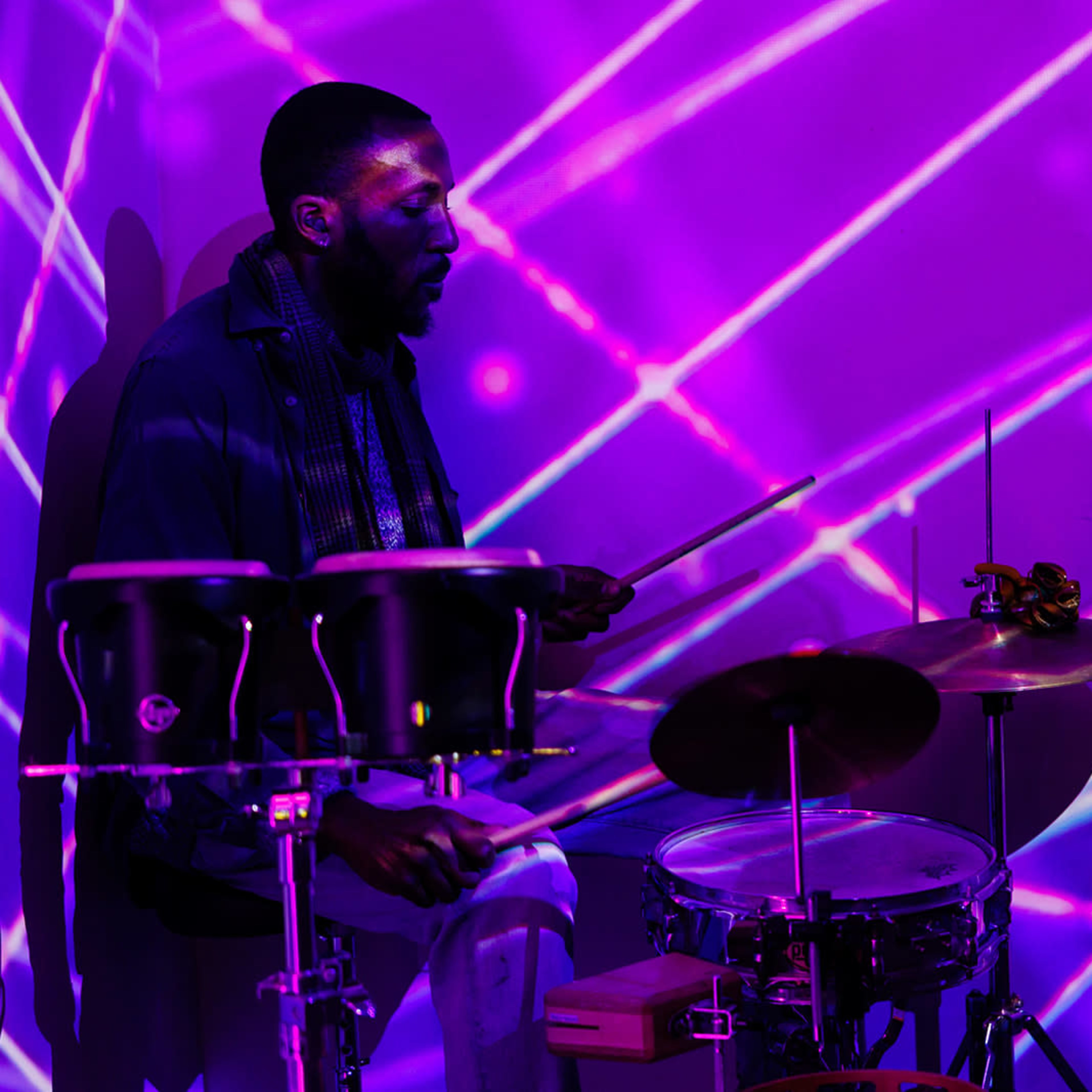 A musician plays the drums against a colorful backdrop of intersecting light beams.