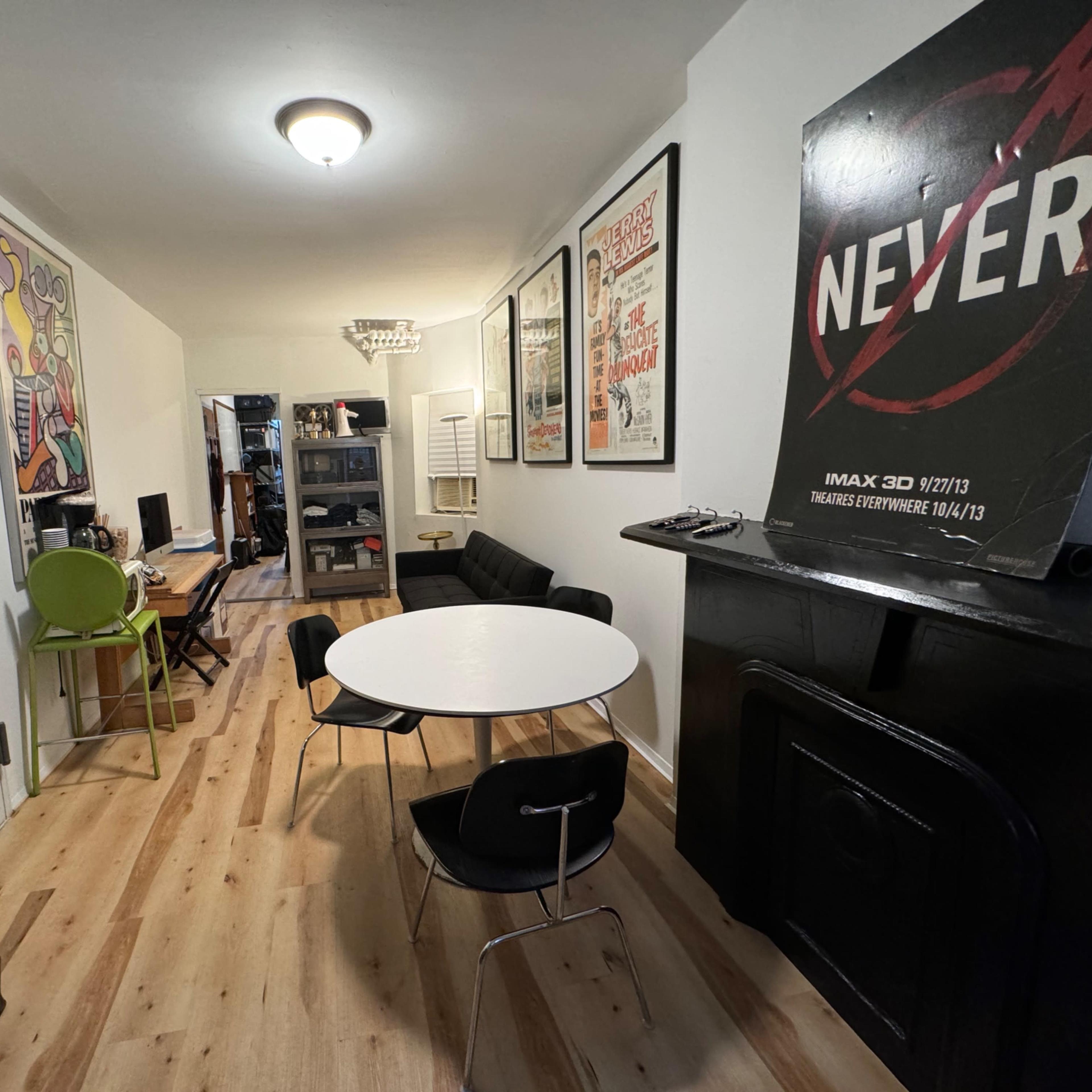 A narrow room with light-colored wooden flooring, featuring a round white dining table surrounded by chairs, modern artwork on the walls, and a black fireplace.
