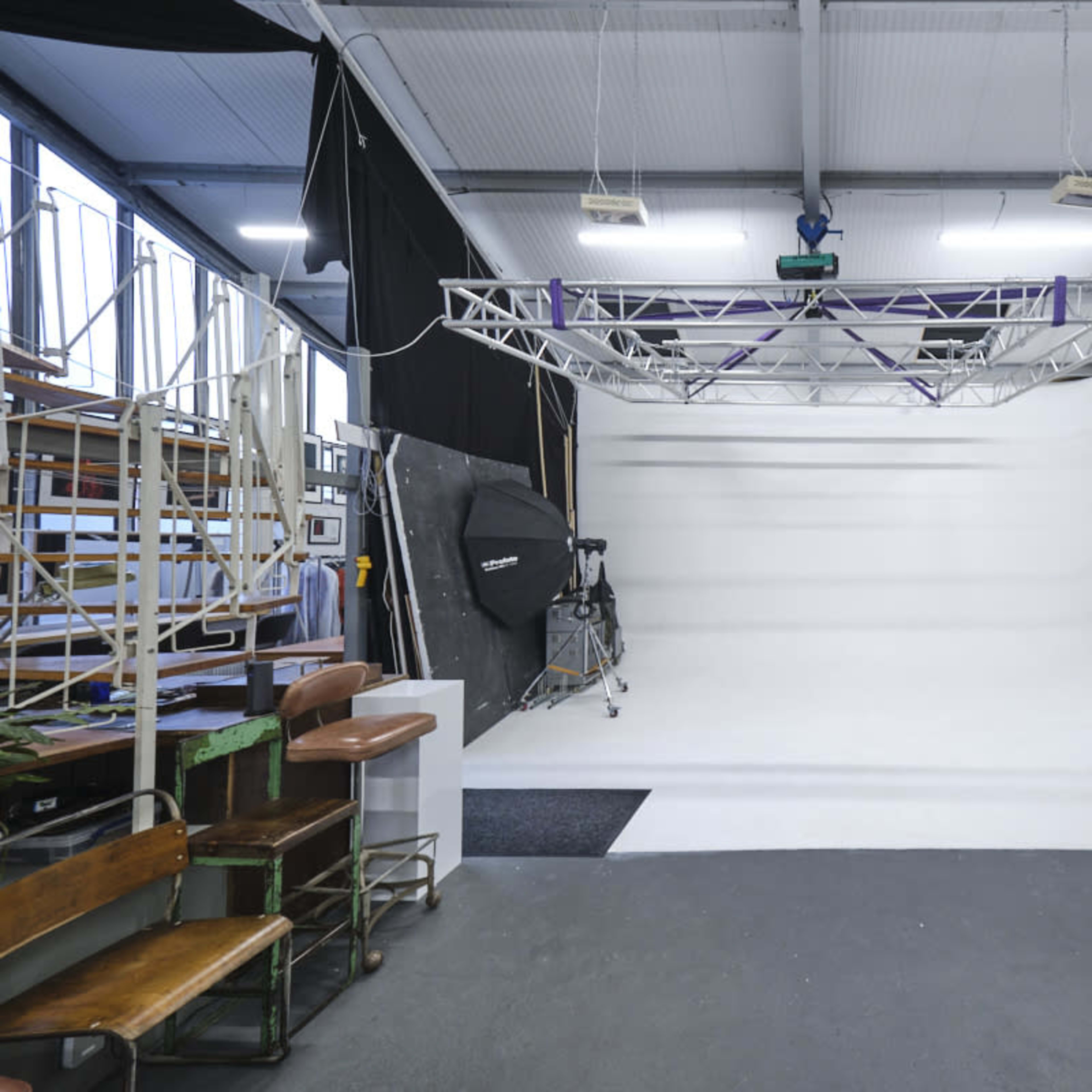 A spacious photography studio with a white background, a lighting rig overhead, and various equipment along the sides.