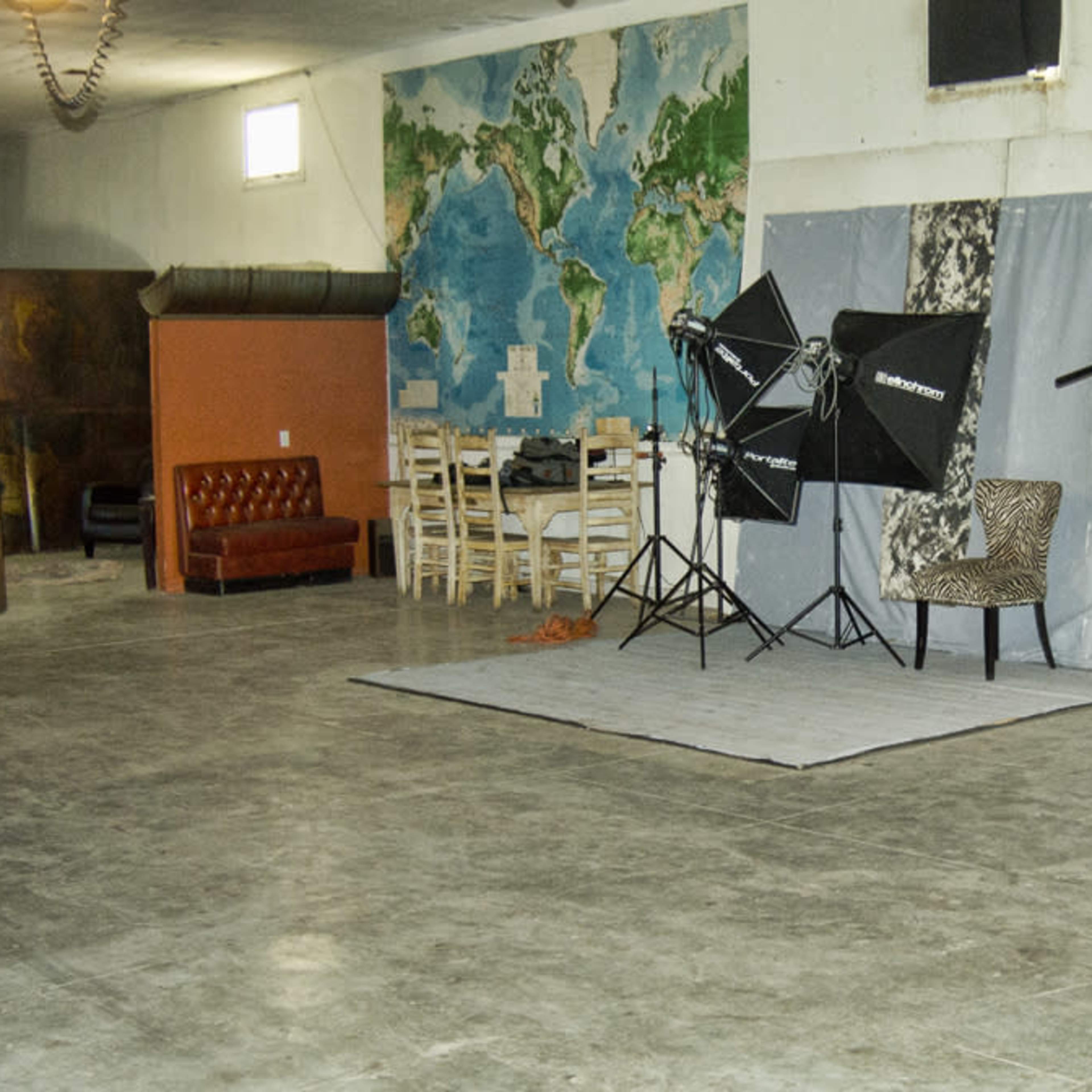 A spacious studio with a world map mural, photography lighting equipment, a patterned chair, and a wooden dining table with chairs.