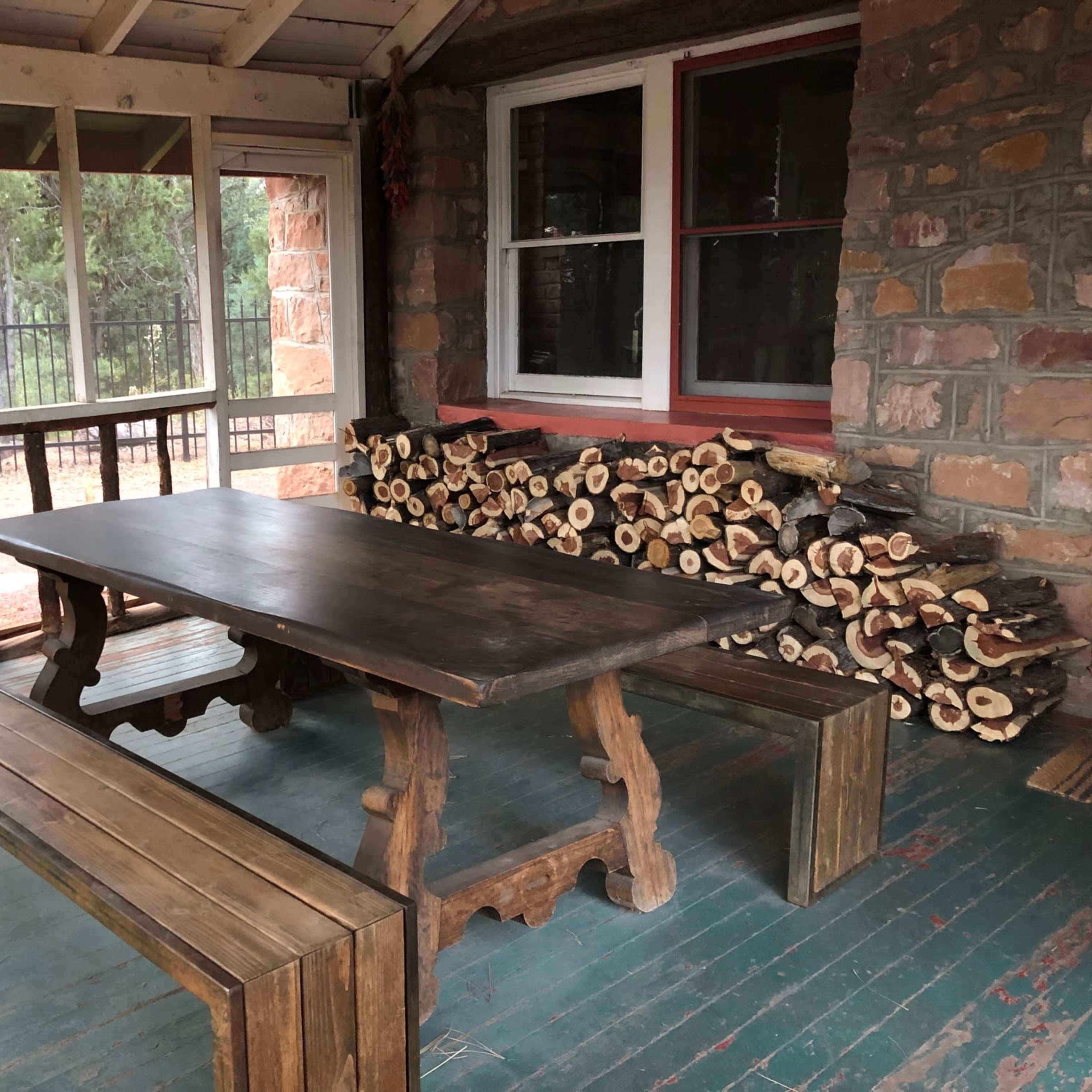 A wooden table and benches are positioned on a porch next to a stack of firewood against a stone wall.