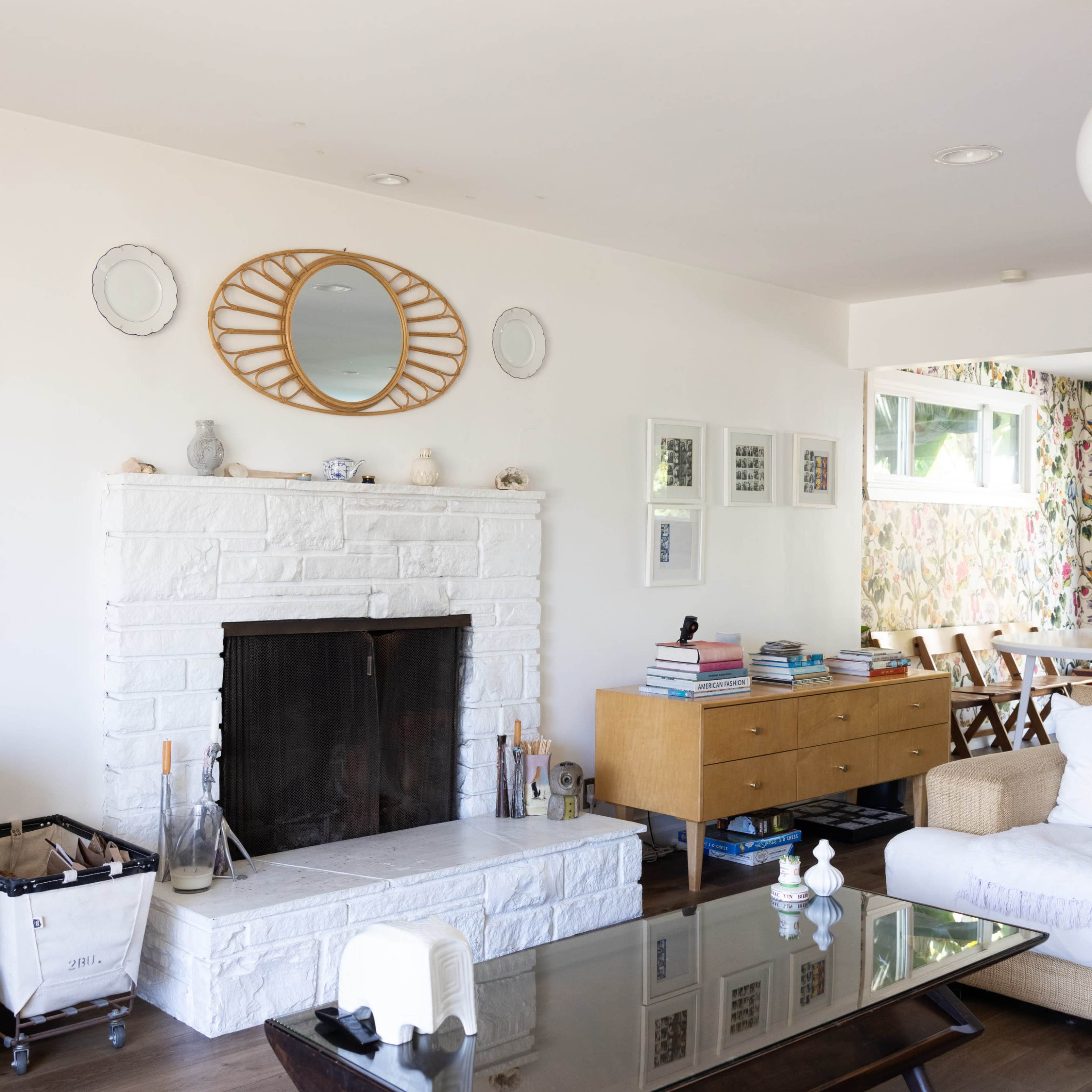 A spacious living room featuring a white stone fireplace, a round mirror above it, a wooden sideboard, and a coffee table in front with various items.