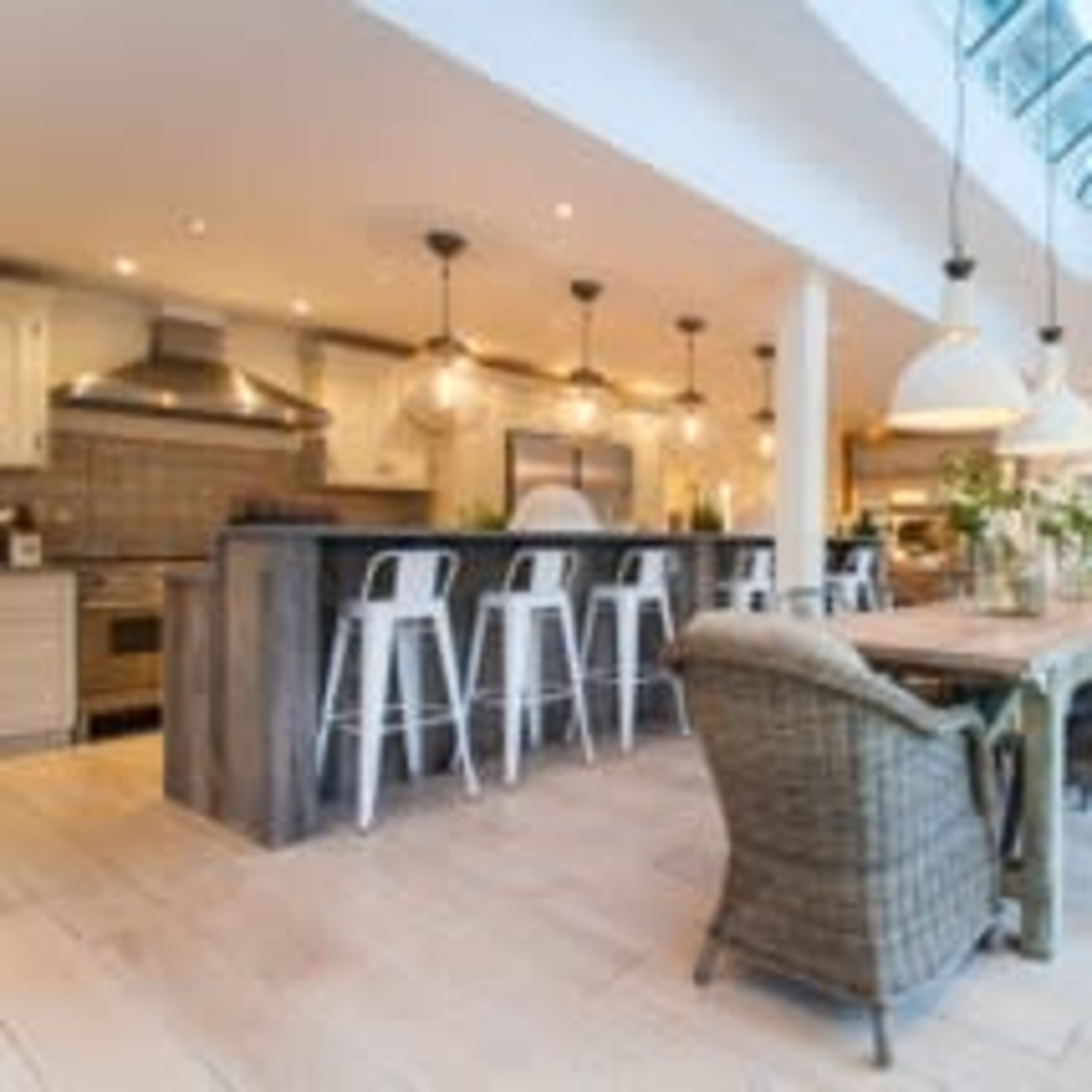 A spacious kitchen area features a large wooden dining table, white bar stools, and modern lighting fixtures with a large skylight overhead.