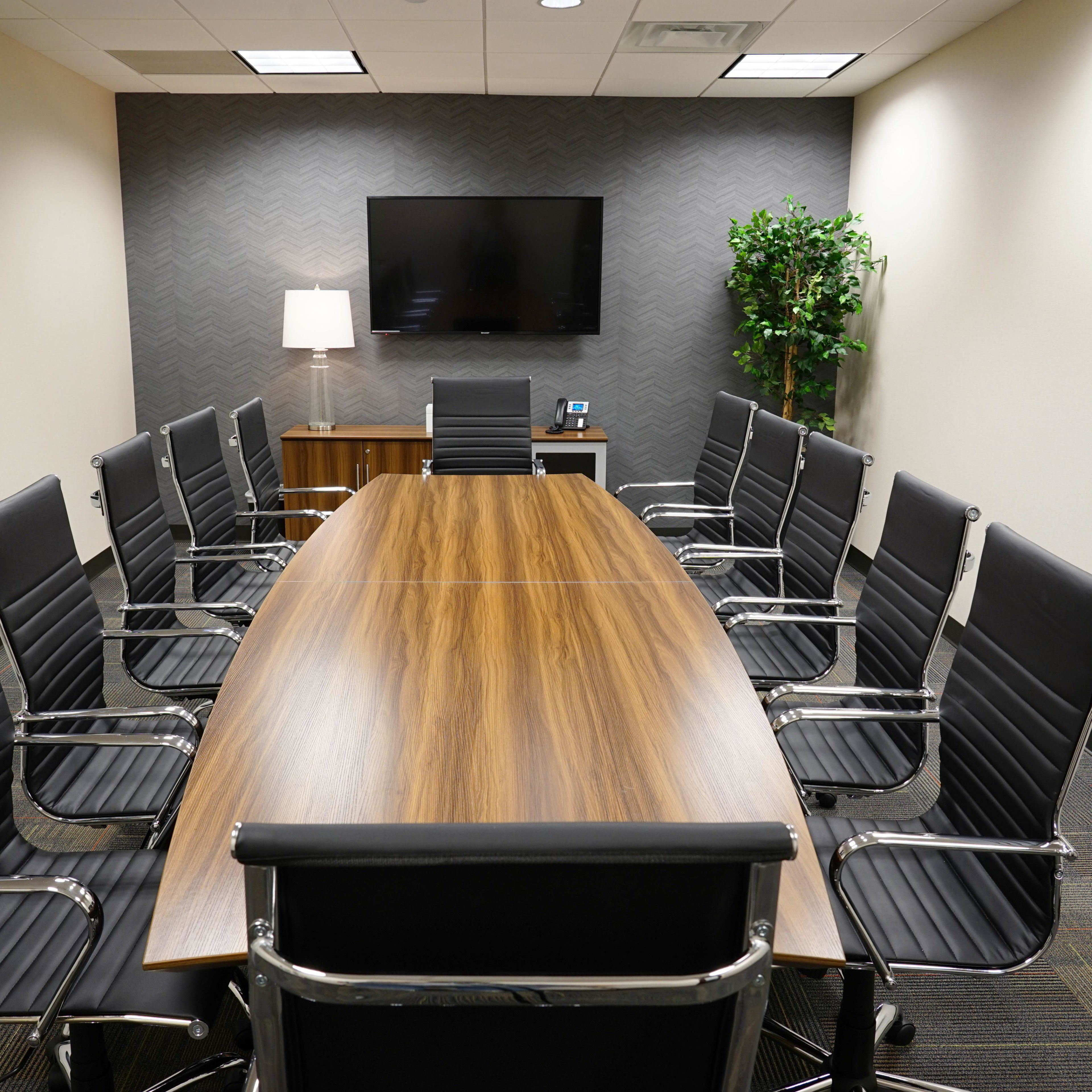 A modern conference room features a large wooden table surrounded by black leather office chairs, with a television and a plant positioned in the background.