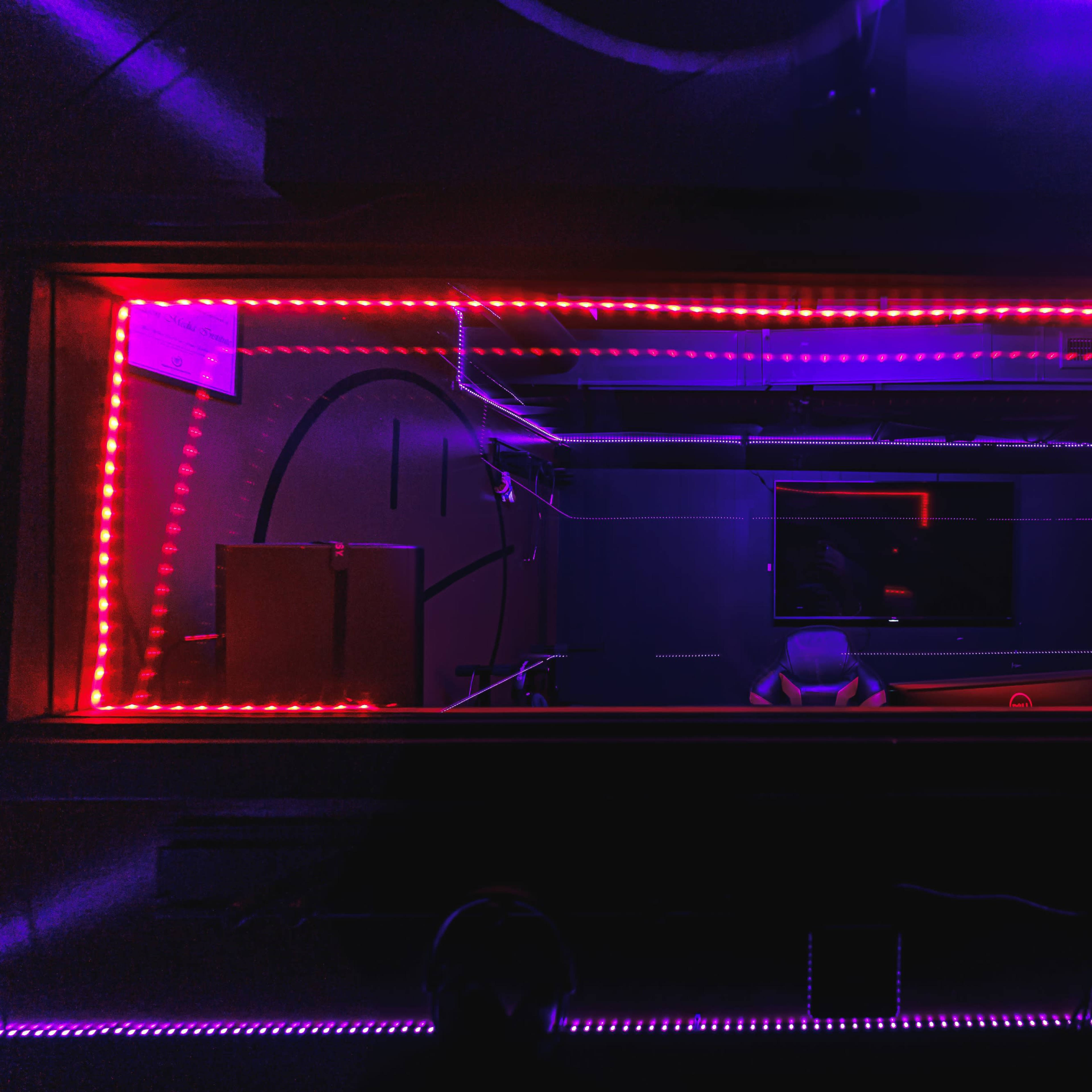 A dimly lit recording studio enclosed in glass, featuring red LED lights, audio equipment, and a chair facing a monitor.