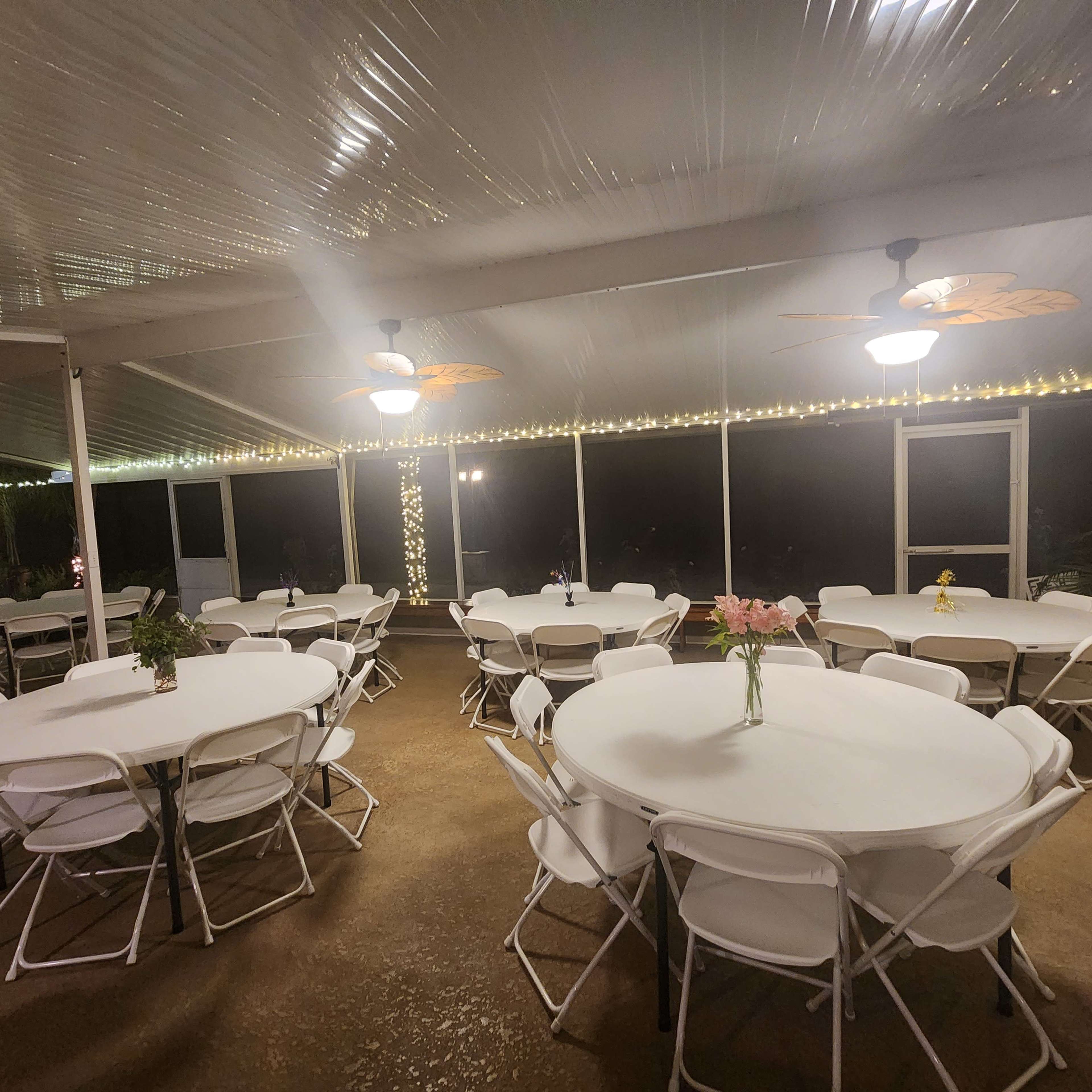 A brightly lit outdoor event space with several round tables arranged under a clear roof, adorned with string lights.