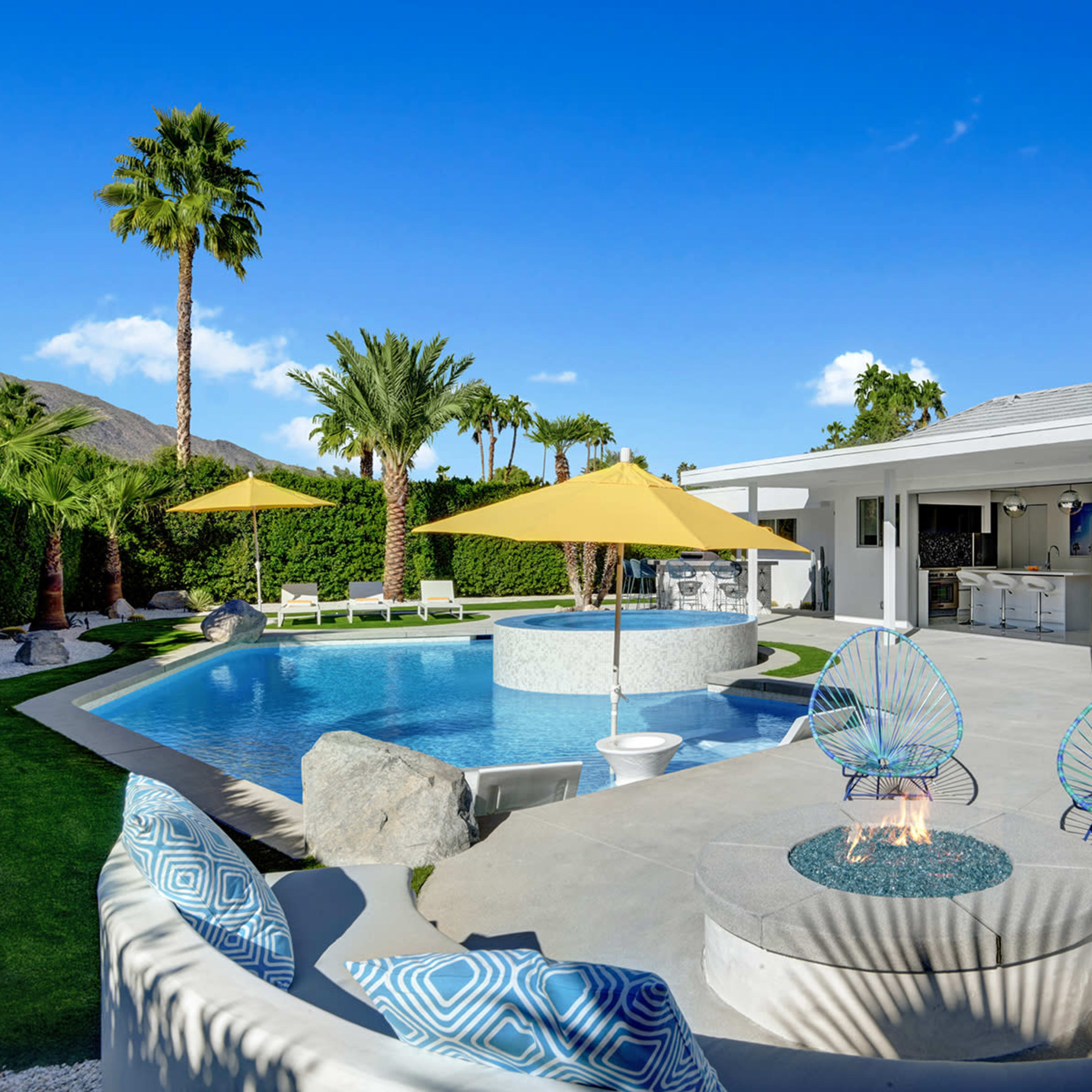 A modern backyard features a pool with lounge chairs, vibrant umbrellas, and a patio area with a fire pit surrounded by colorful chairs.