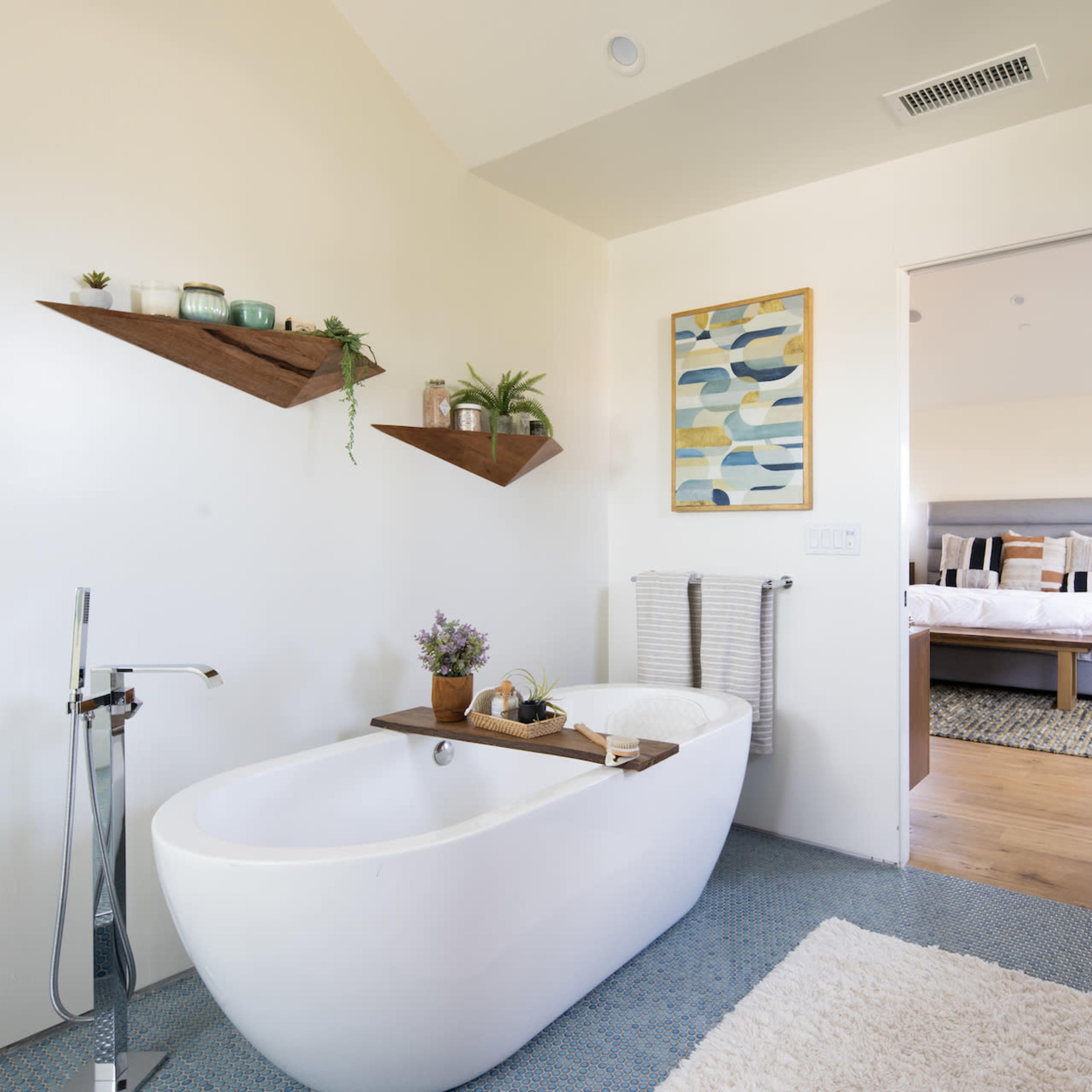 A modern bathroom with a freestanding bathtub, a wooden shelf with plants, and an open doorway leading to a bedroom.