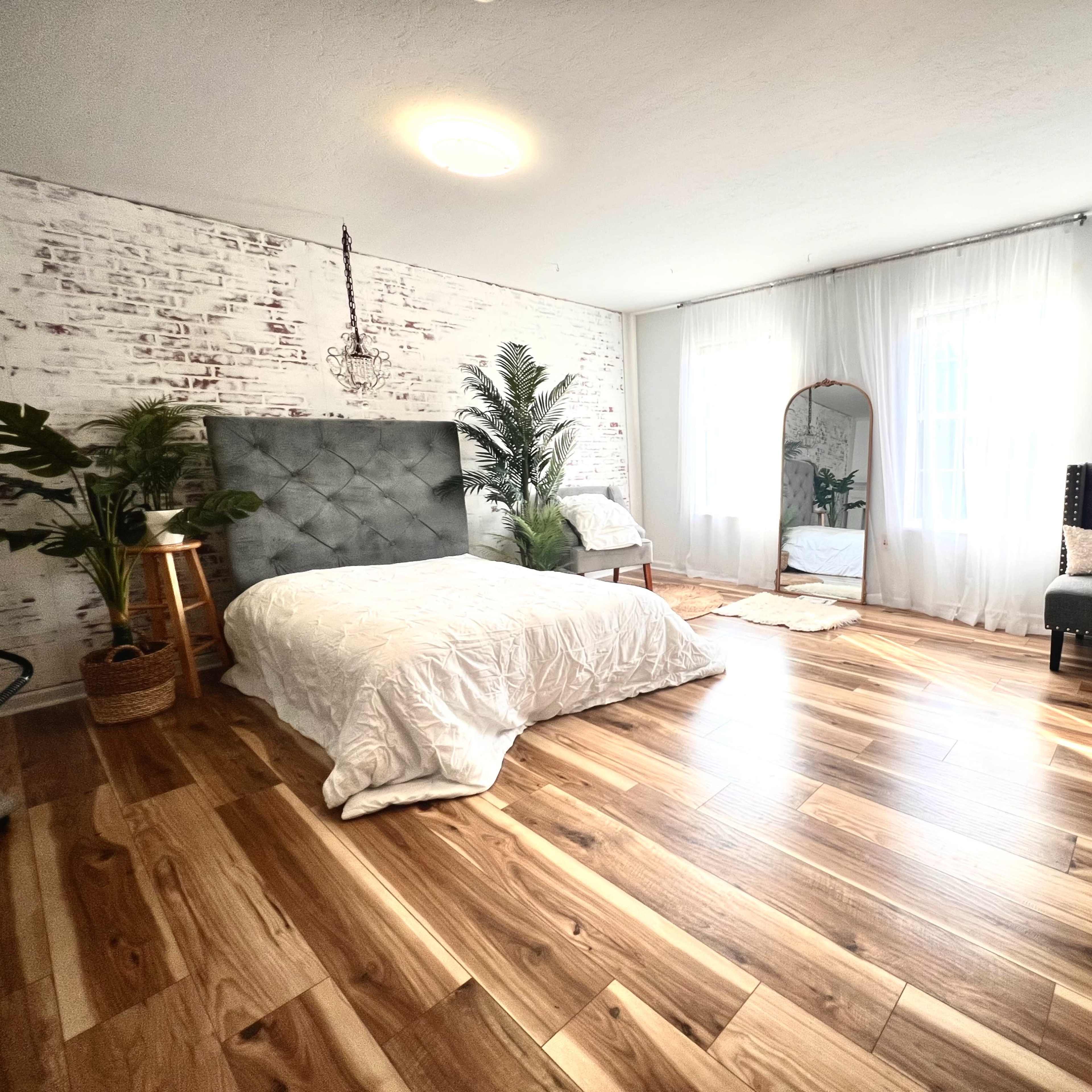 The room features a bed with a gray upholstered headboard, a large mirror, potted plants, and hardwood flooring, illuminated by natural light from two windows.