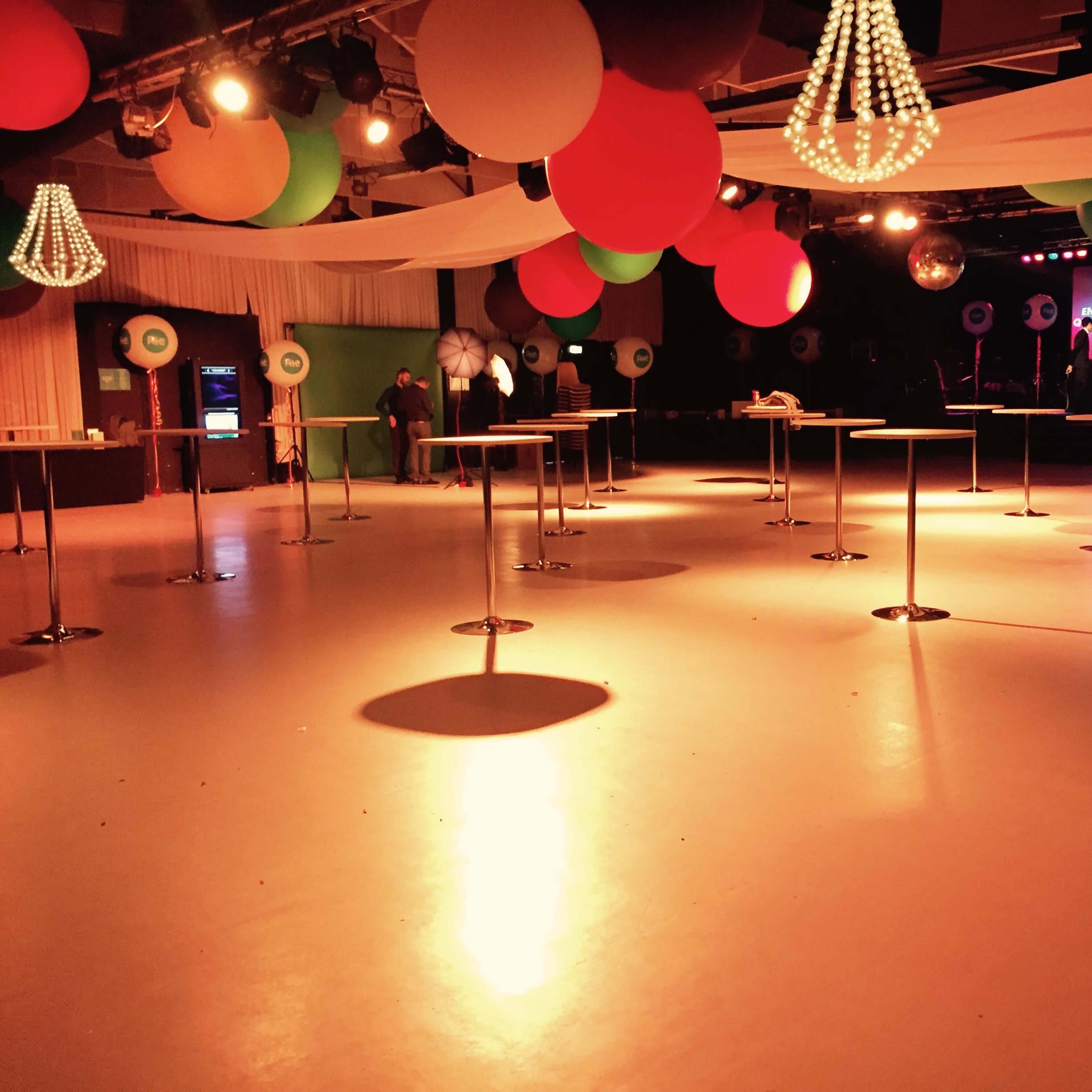 An empty event space with round tables, colorful hanging decorations, and a stage at the back.