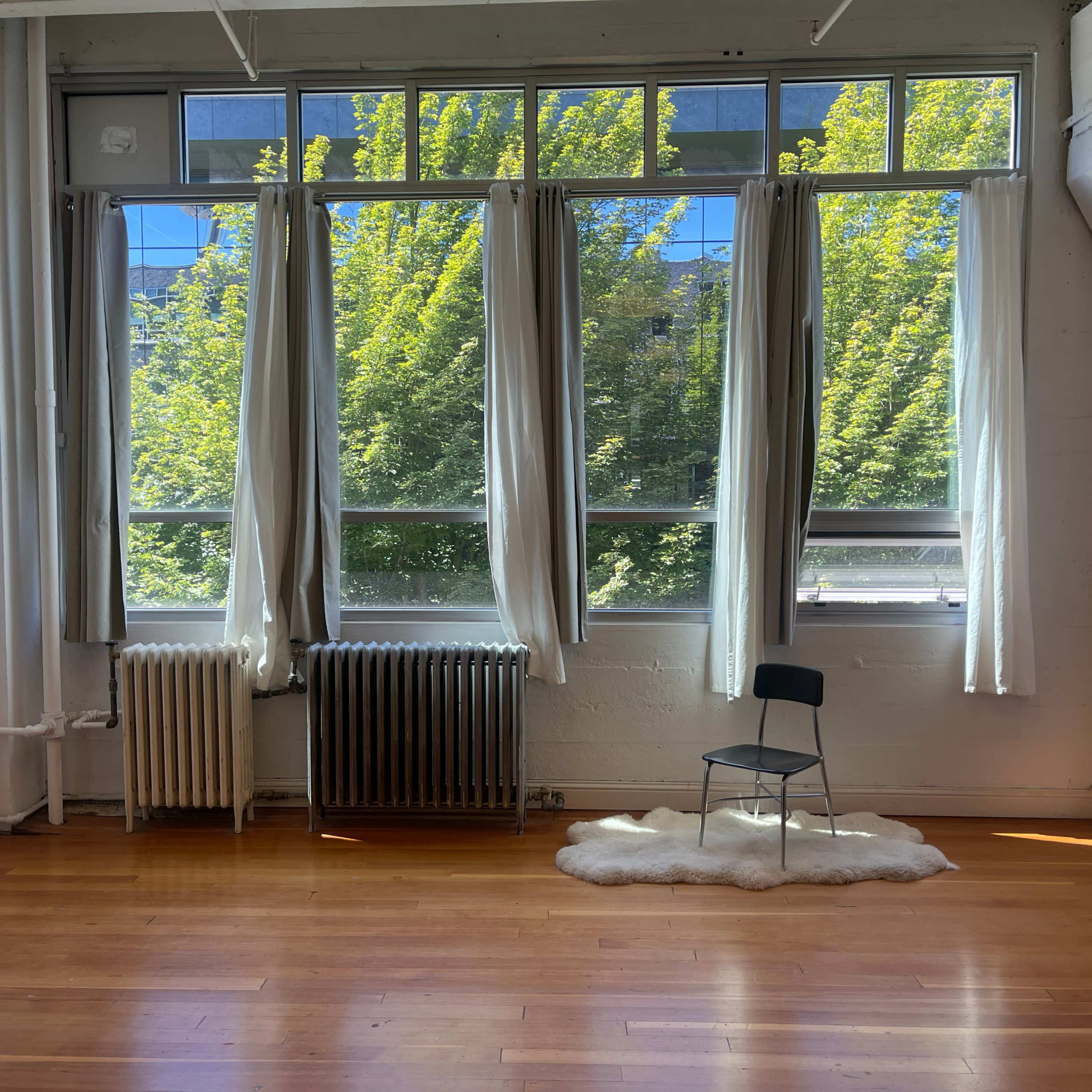 A room features large windows with sheer curtains, a lone chair on a white rug, and a radiator beneath the windows, revealing green trees outside.