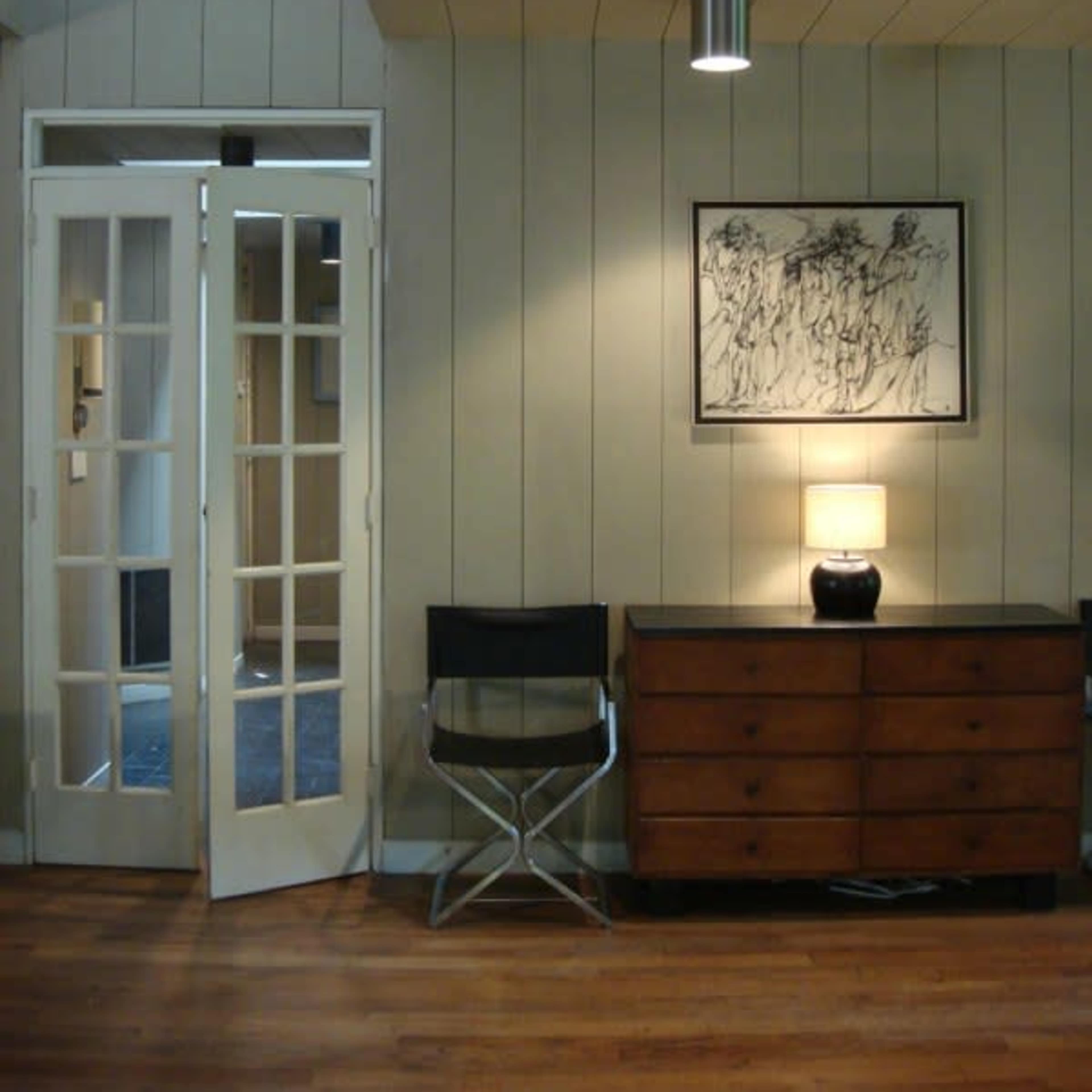 A room with a wooden floor, white-paneled walls, a framed artwork, a lamp on a dresser, and a folding chair next to double doors.