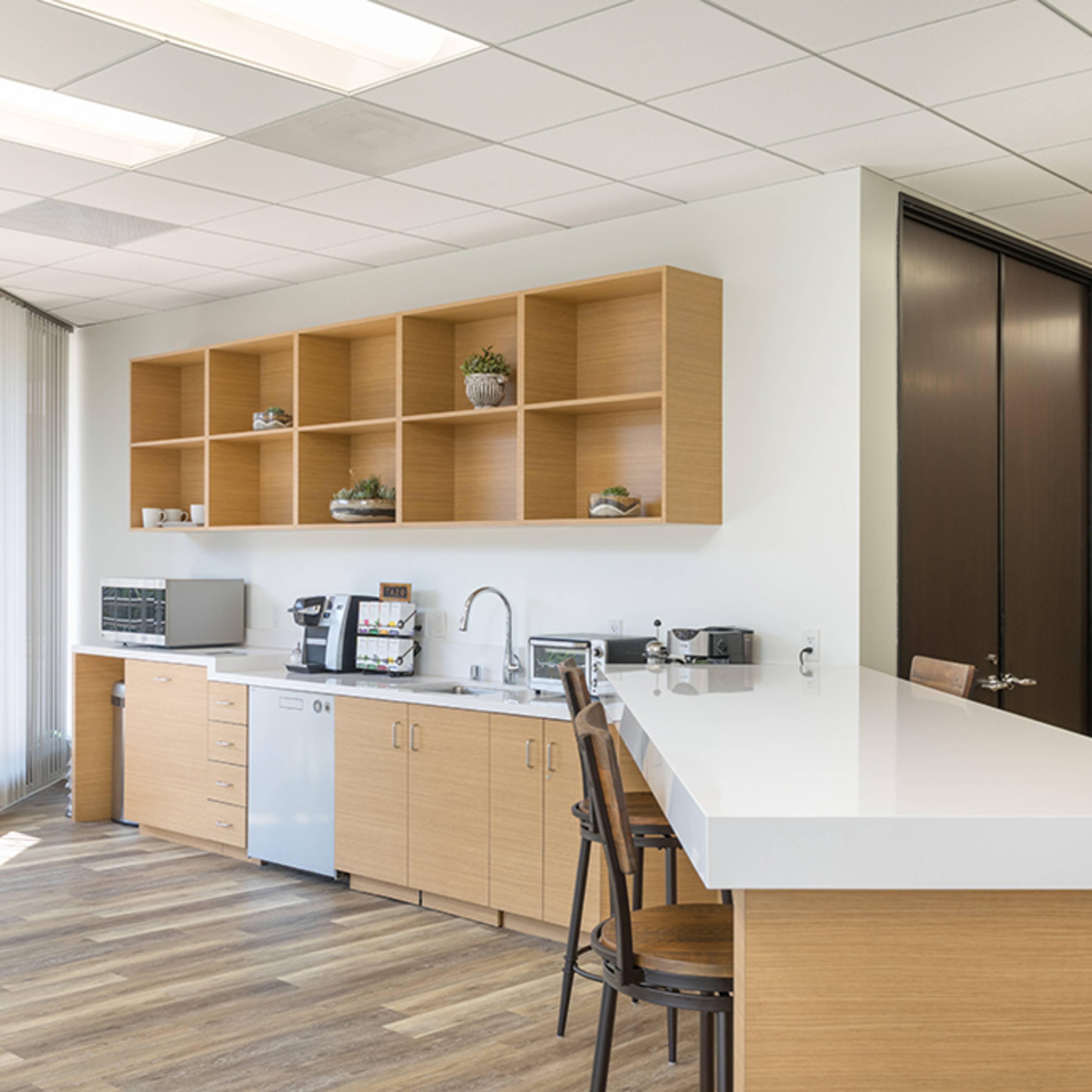 A modern office kitchenette with wood cabinetry, a white countertop, and various appliances neatly arranged.